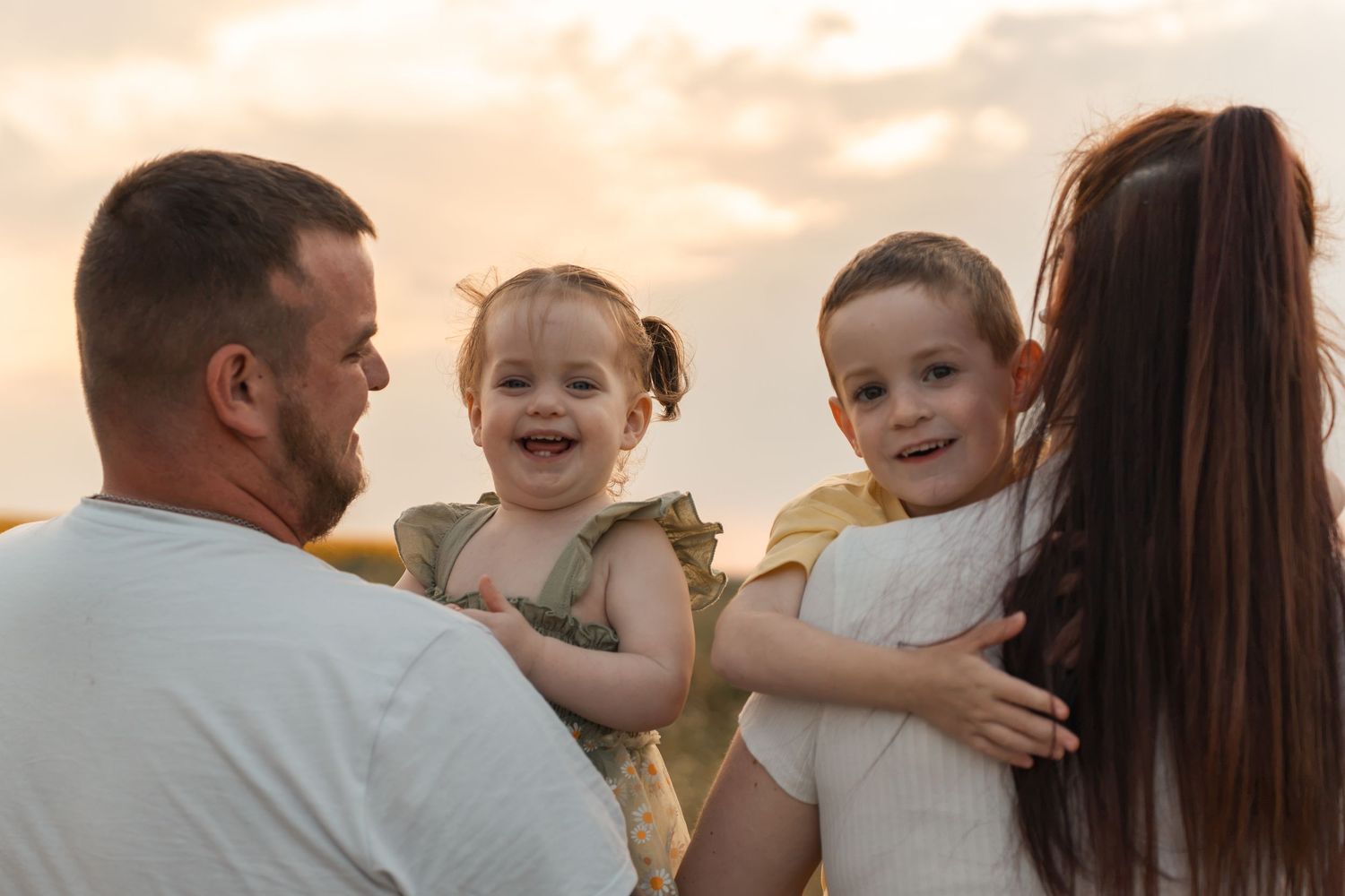 Happy family with two smiling toddlers held by parents at sunset in an outdoor field setting.