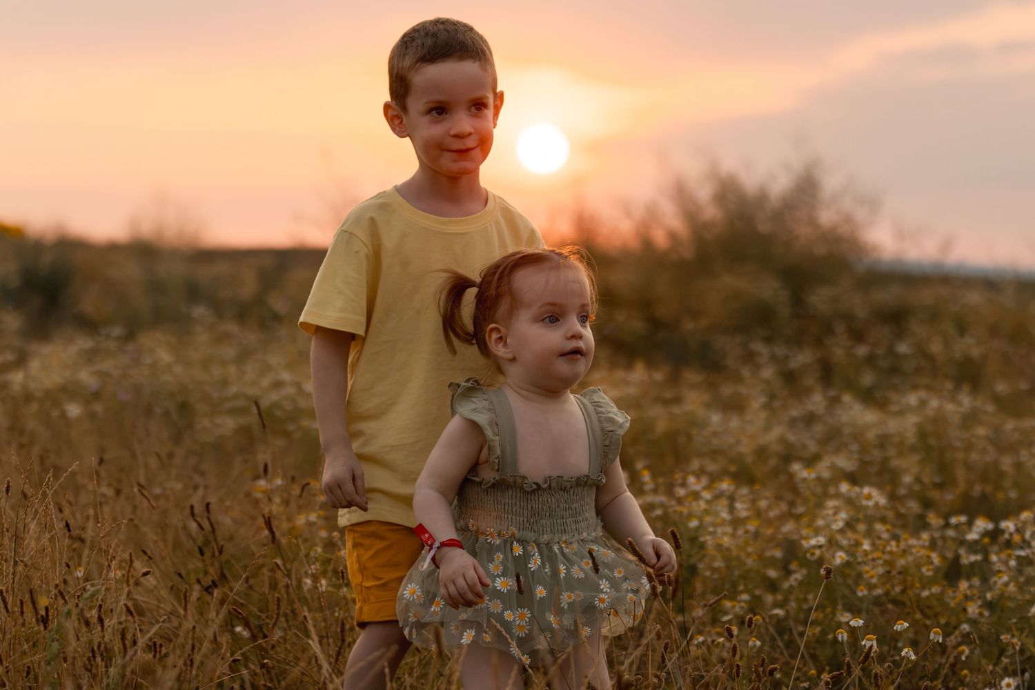 Two young children pose in a golden field at sunset, older boy in yellow shirt with toddler girl in floral dress.