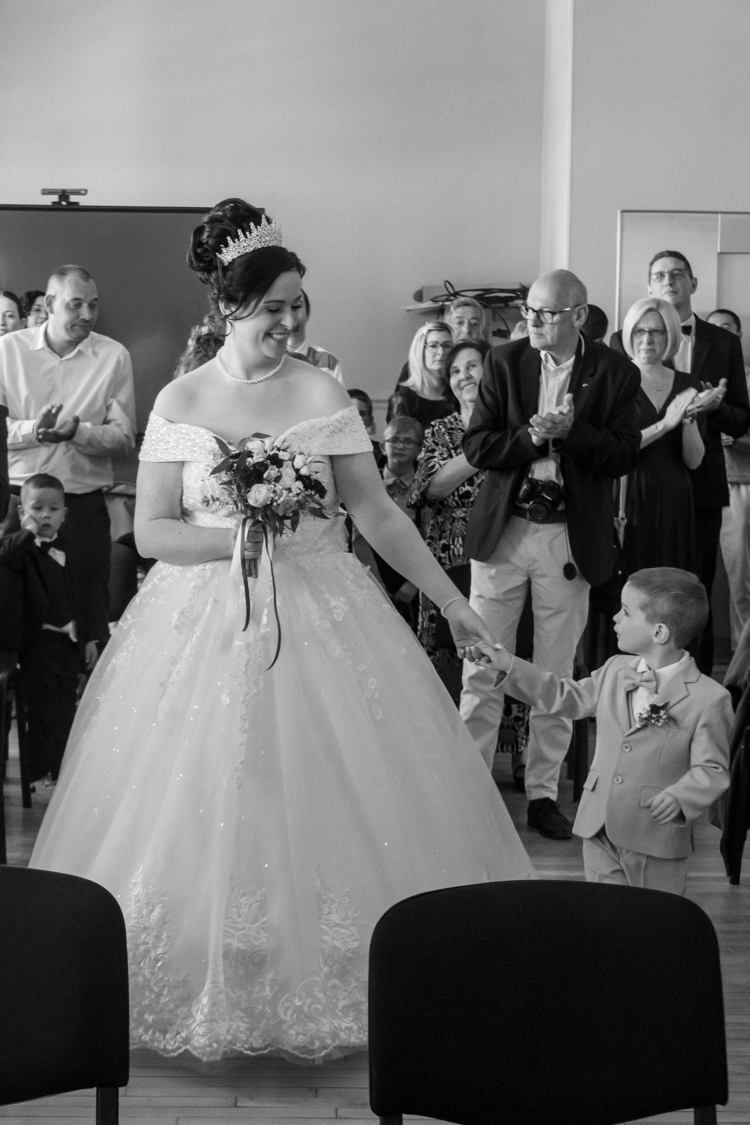 Black and white photo of a smiling bride in a ballgown holding hands with a young boy at a wedding ceremony.