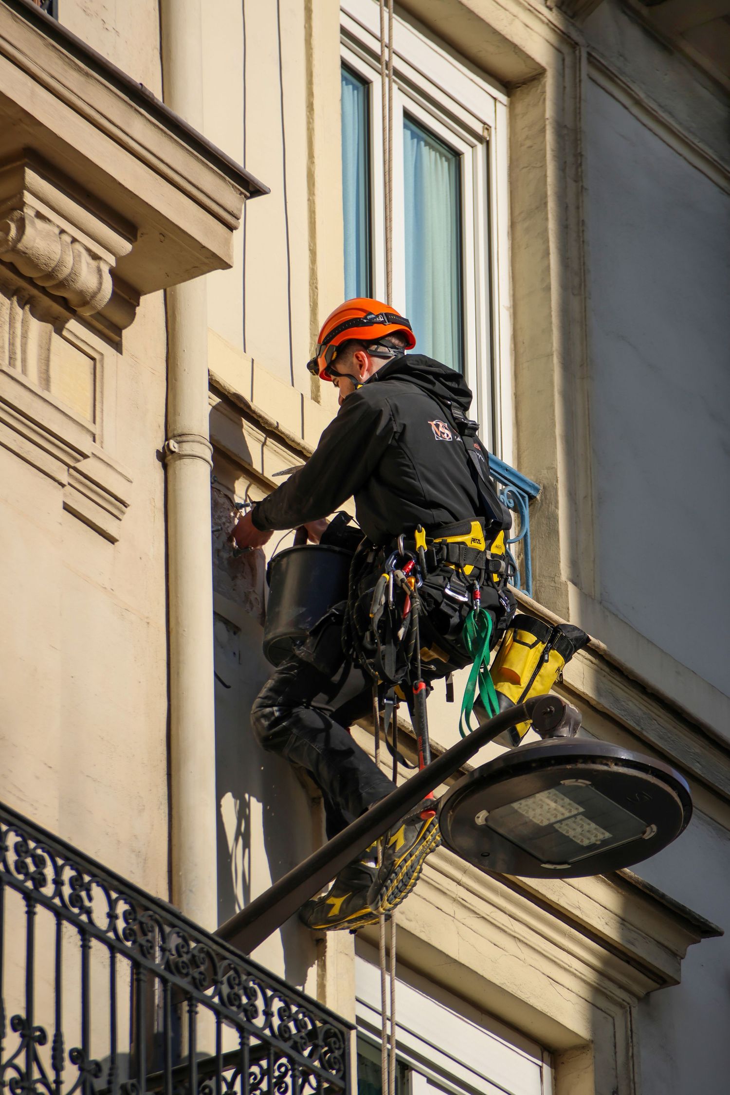 Worker in orange helmet and safety gear performing maintenance on a classic stone building facade from a platform.