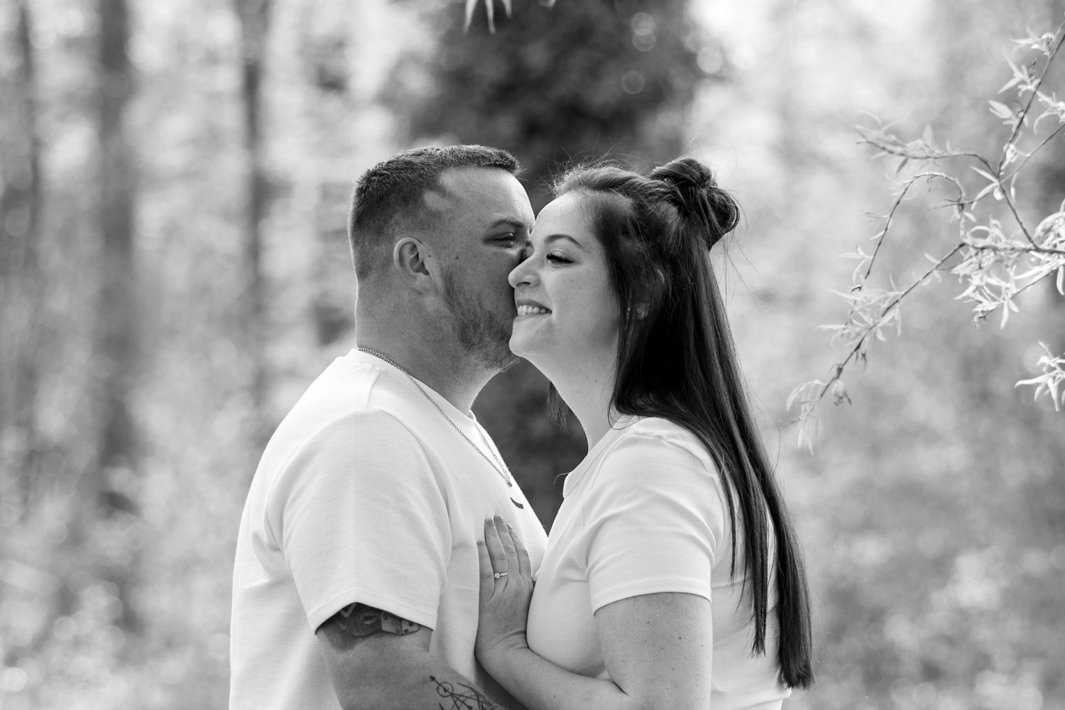 Black and white photo of a couple embracing and smiling in a forest setting, man kissing womans cheek tenderly.