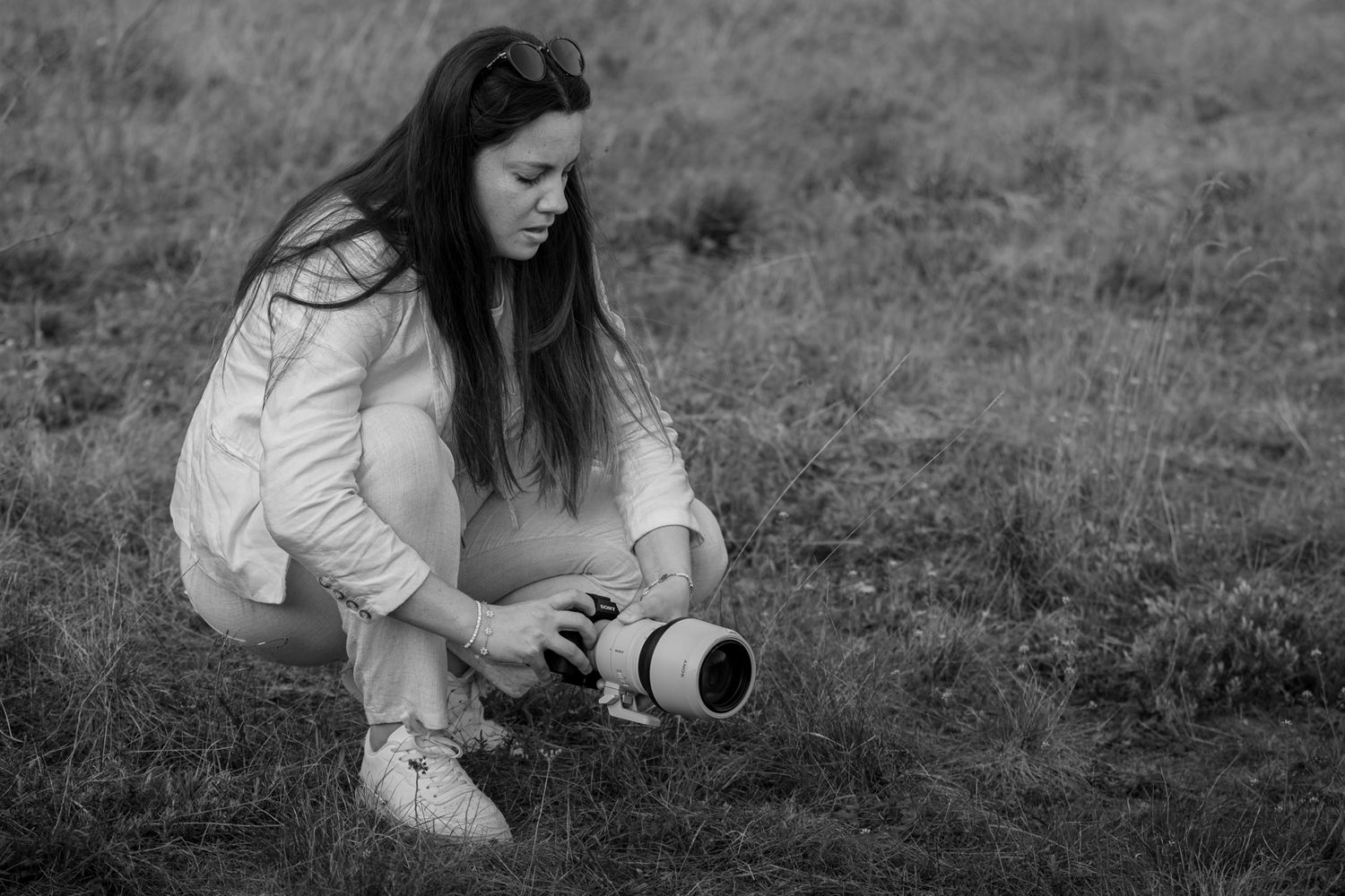 Black and white photo of a young woman crouching in a field, holding a professional camera with a large lens.