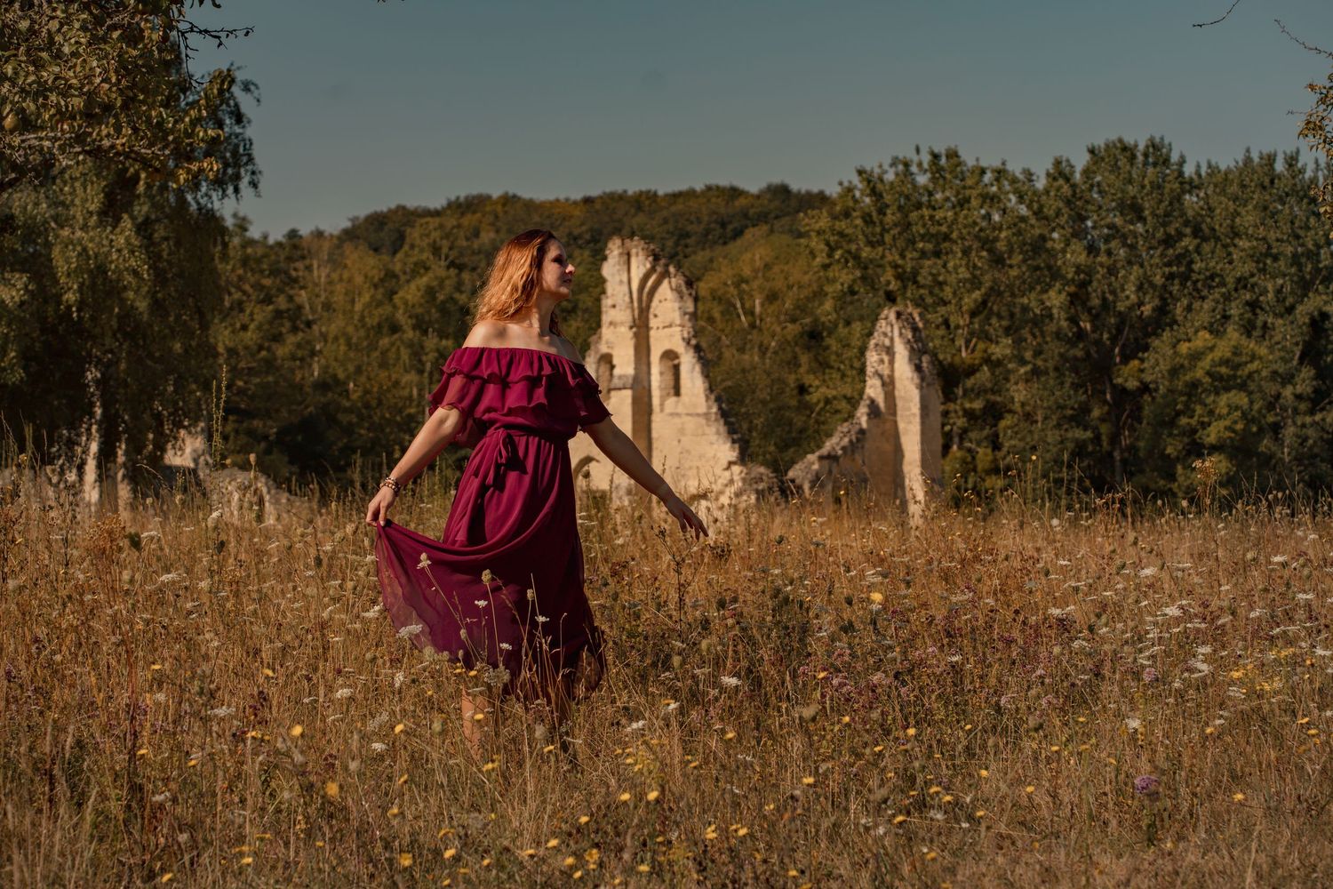 Woman in burgundy dress walking through wildflower field near ancient stone ruins on a sunny day.