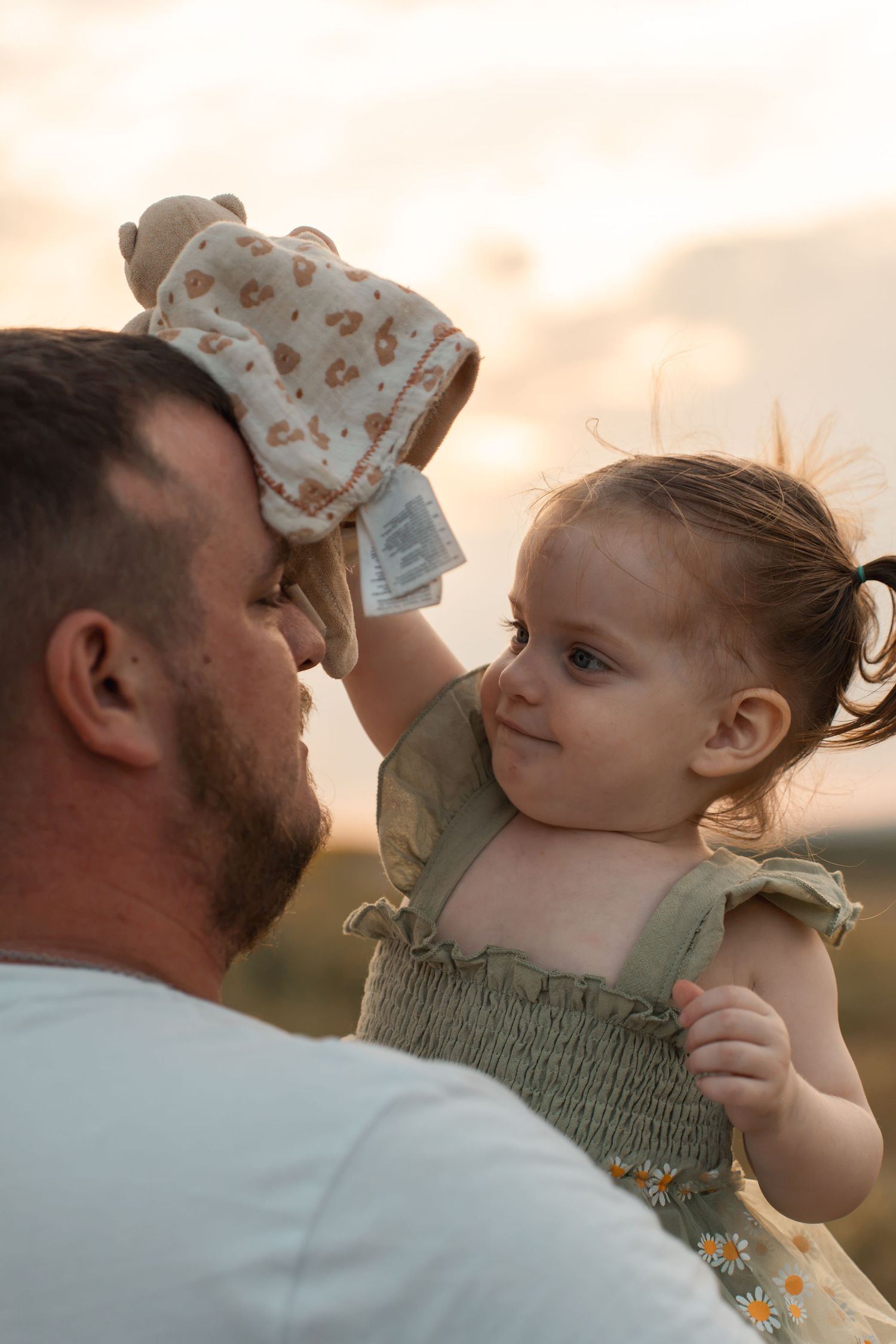 A father holds his toddler daughter at sunset as she playfully covers his face with a stuffed animal toy.
