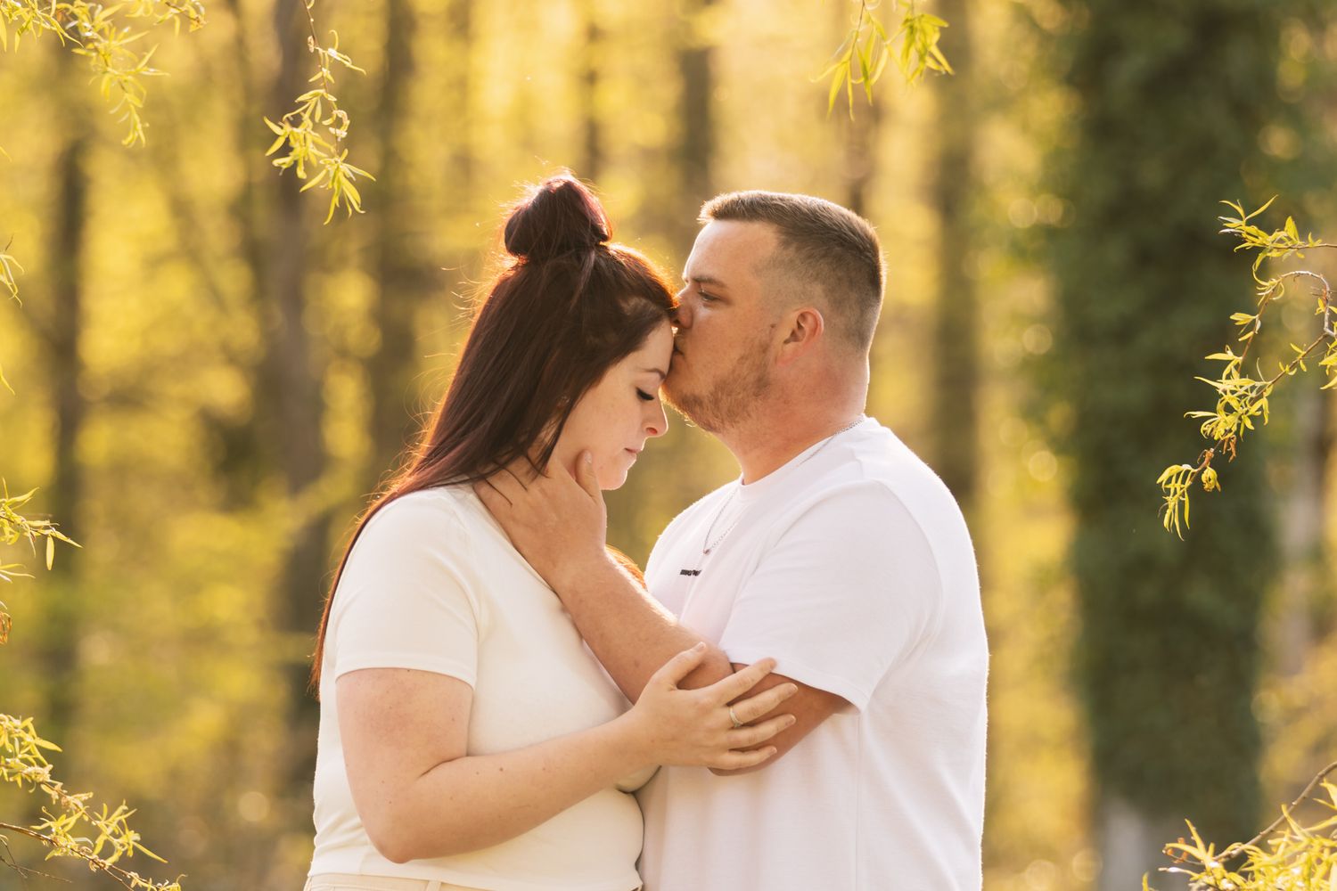 A man in a white shirt kisses a woman's forehead while embracing outdoors among golden autumn trees.