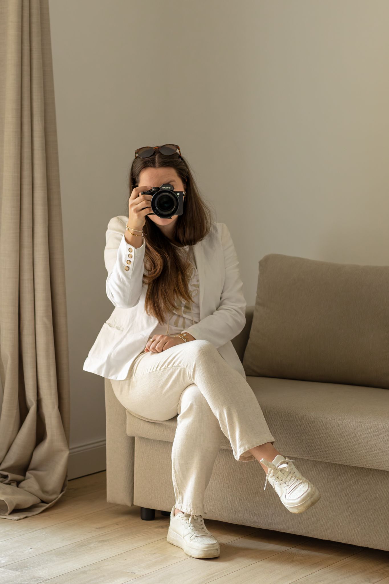 Woman in white outfit sitting on beige sofa, holding DSLR camera up to her face, sunglasses on head, indoor setting.