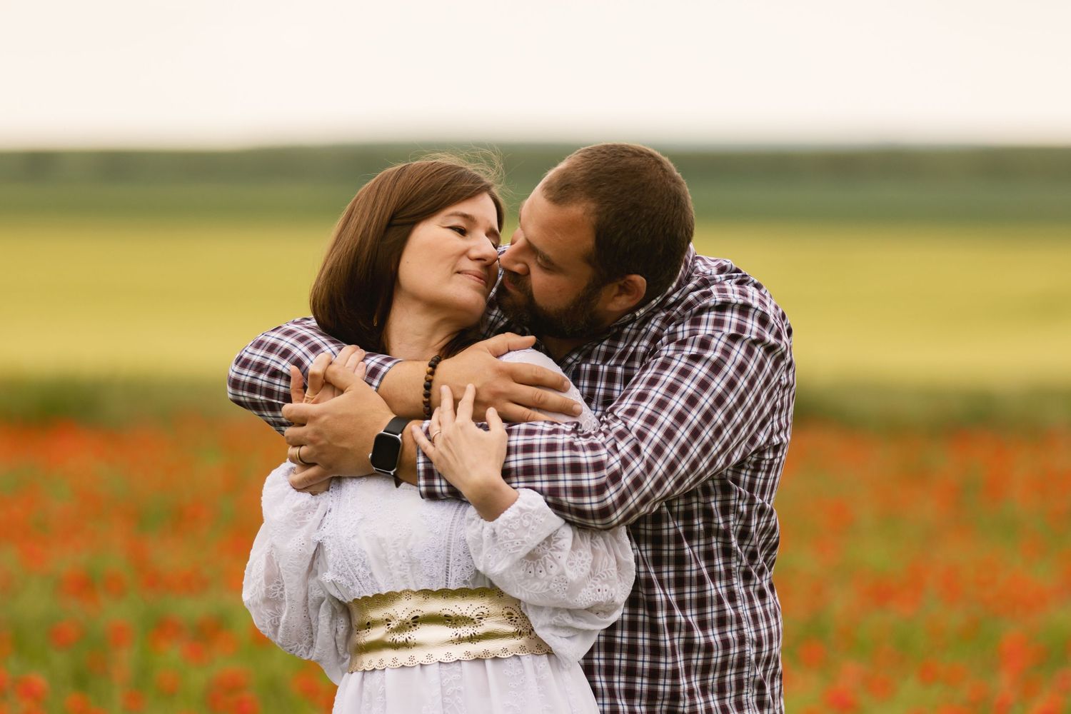 Couple embracing romantically in a vibrant red poppy field, man kissing woman in white dress outdoors.