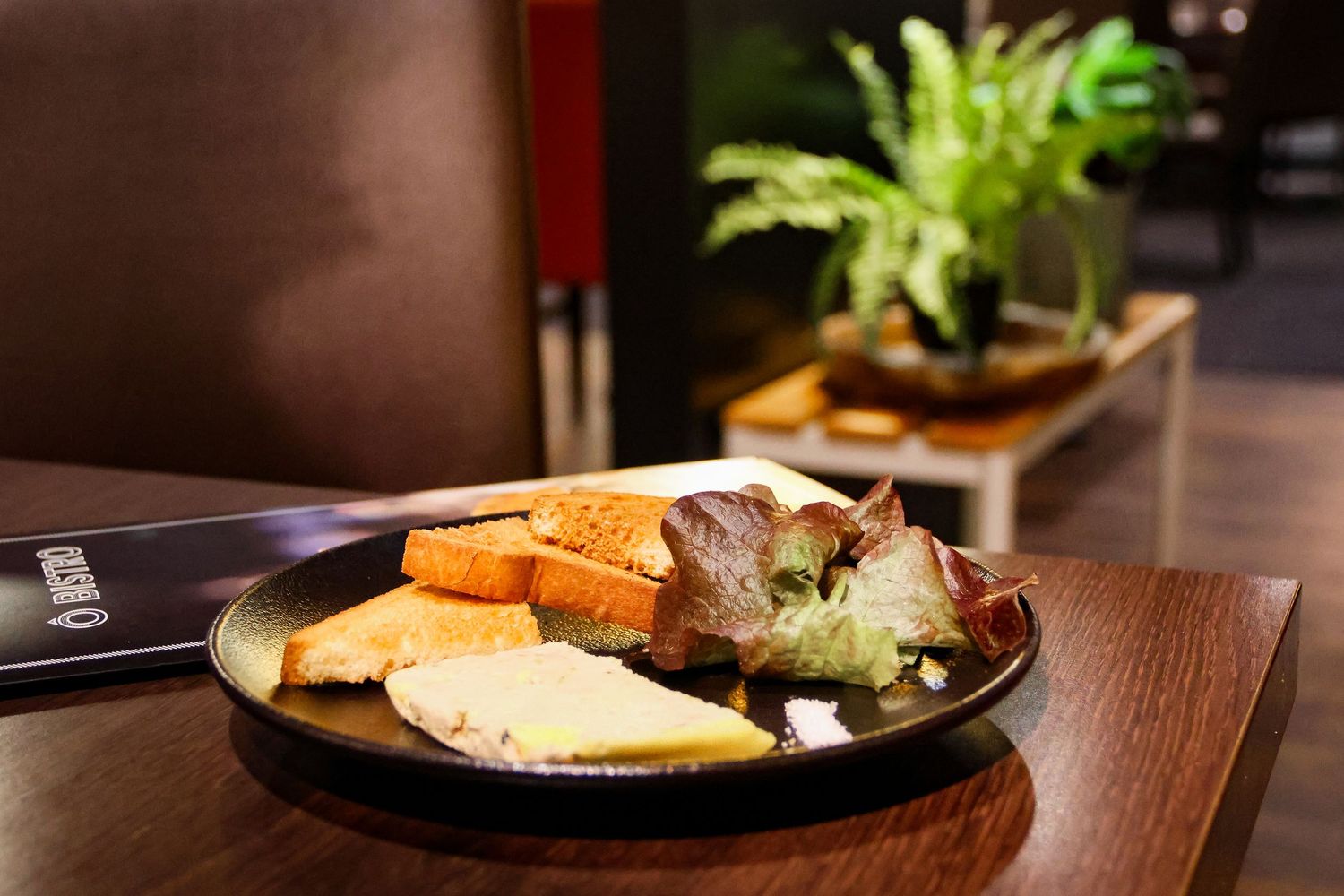 A gourmet cheese and bread platter served on a dark plate on a wooden table with green plants in the background.