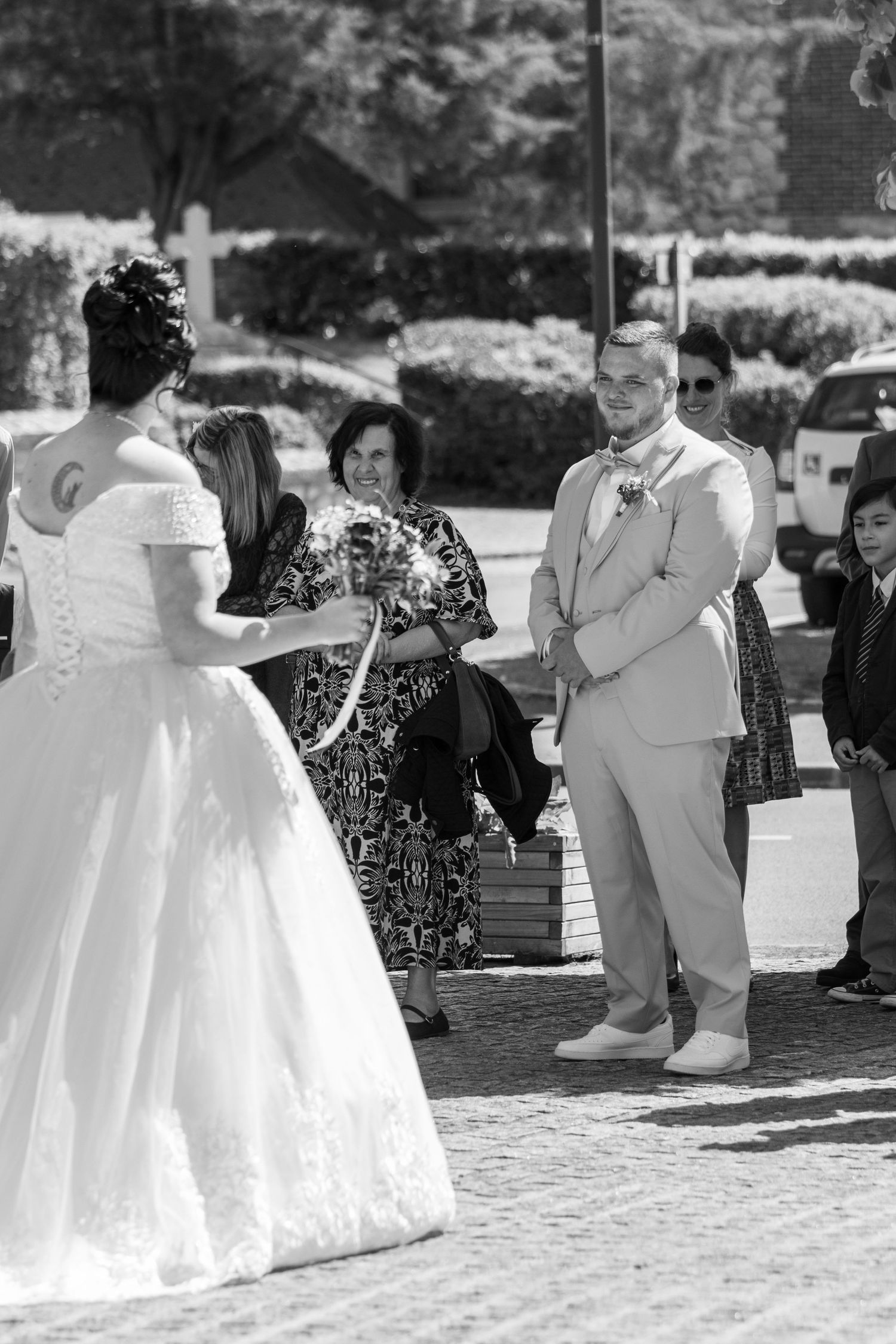 Black and white photo of a bride in a white gown approaching a groom in a tan suit during an outdoor wedding ceremony.