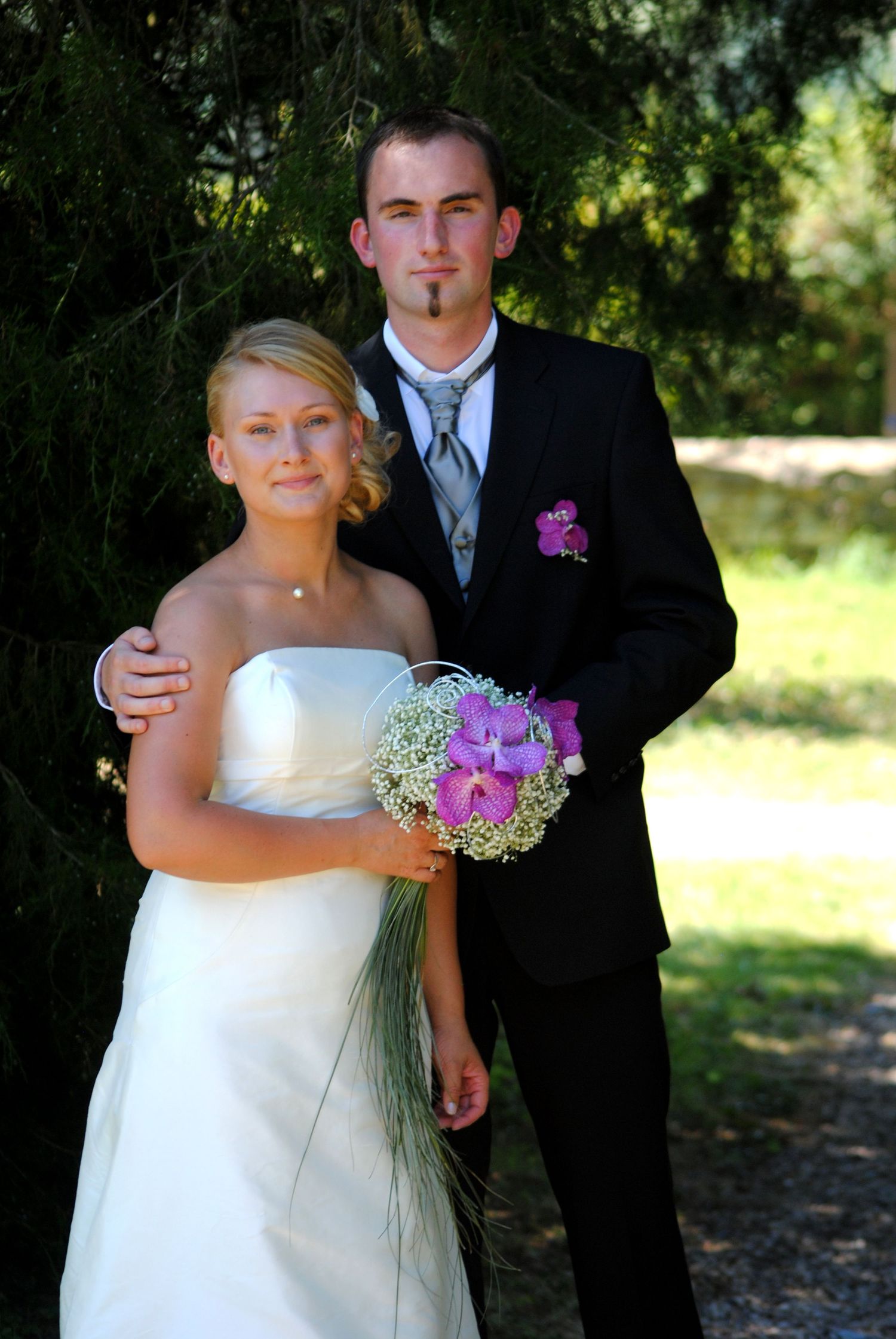 Bride in white strapless gown holding purple floral bouquet stands with groom in black suit outdoors.