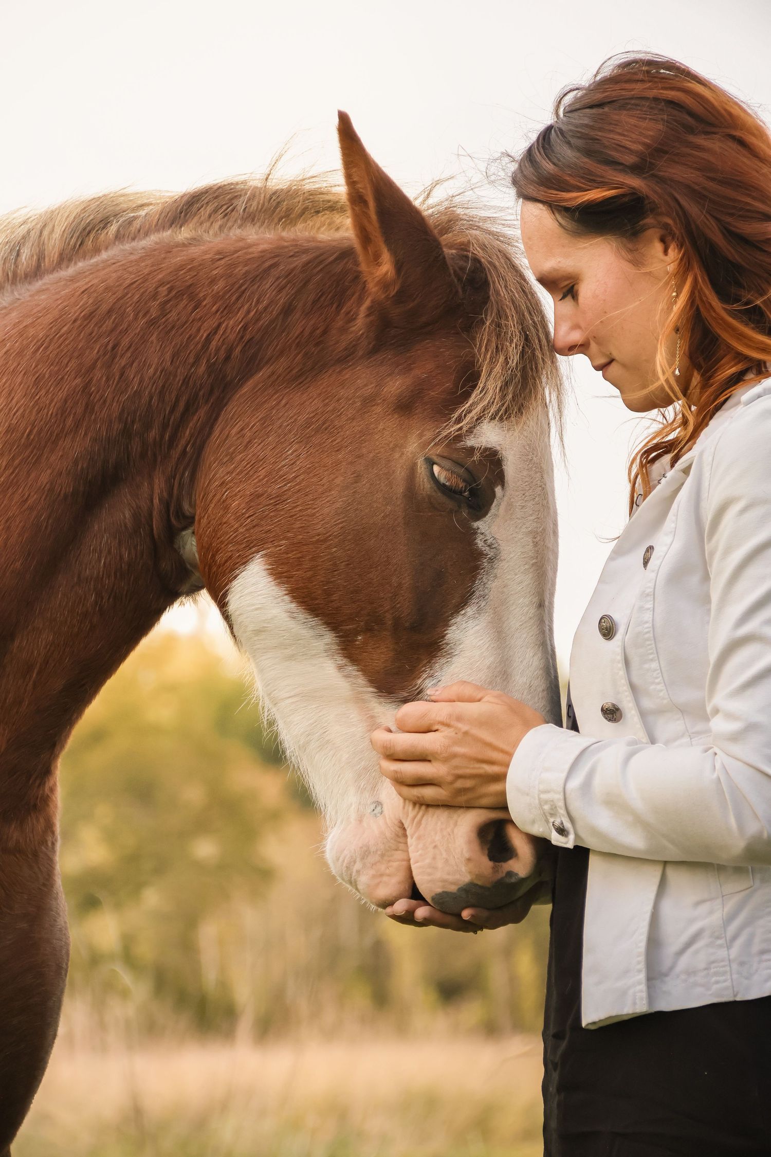 A woman with red hair tenderly holds a chestnut horse's face close to hers in a peaceful outdoor setting.