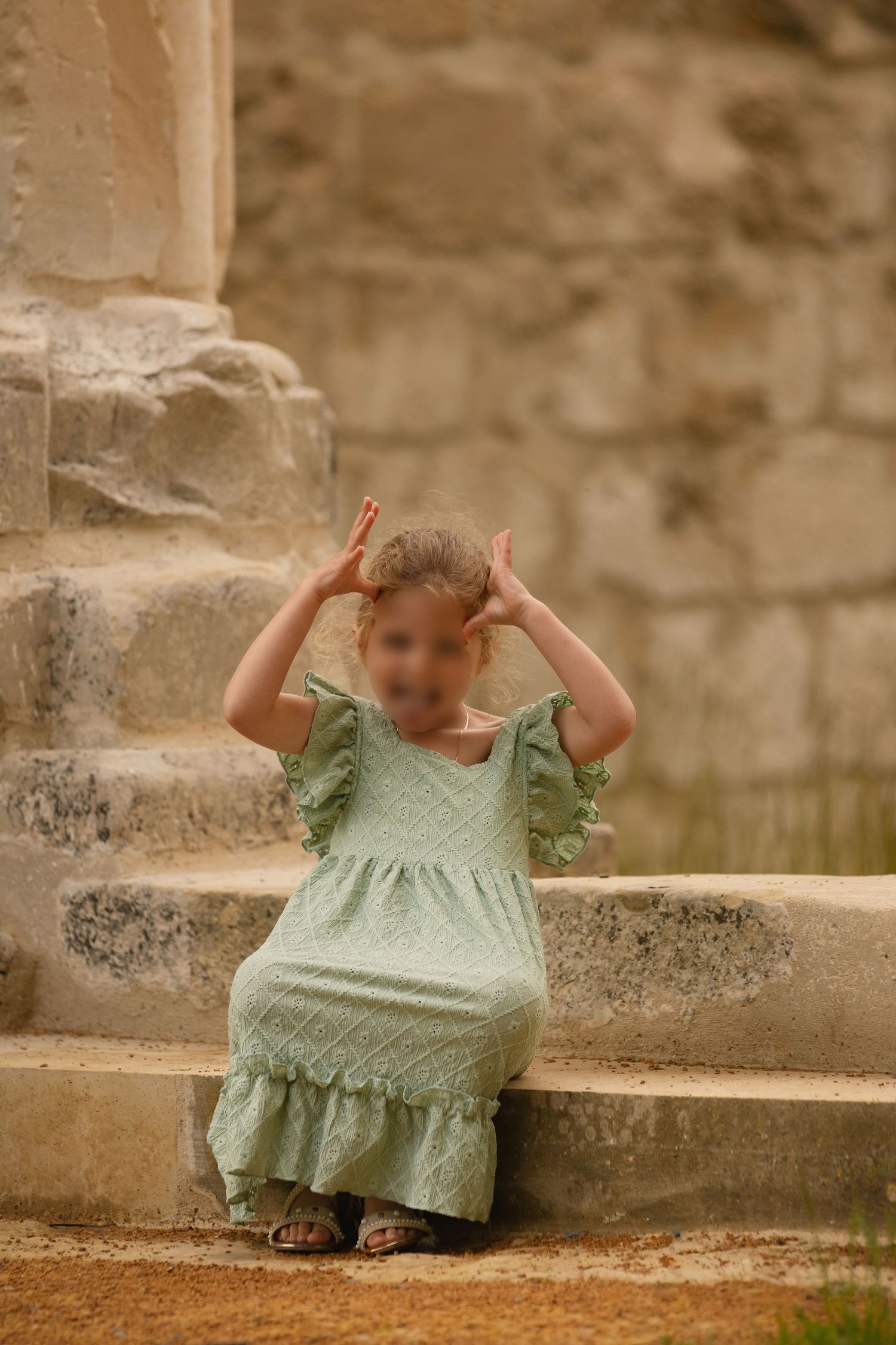 Young girl in mint green ruffle dress sitting on stone steps, playfully holding hands above head outdoors.