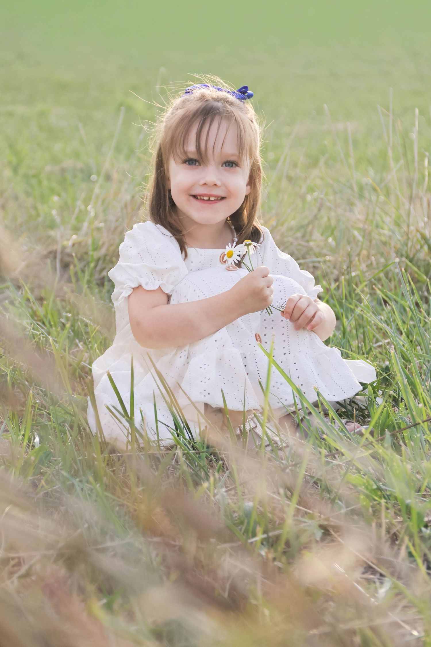 Séance famille lifestyle — fillette souriante en robe blanche dans les herbes hautes tenant un bouquet de marguerites, photographe Soissons