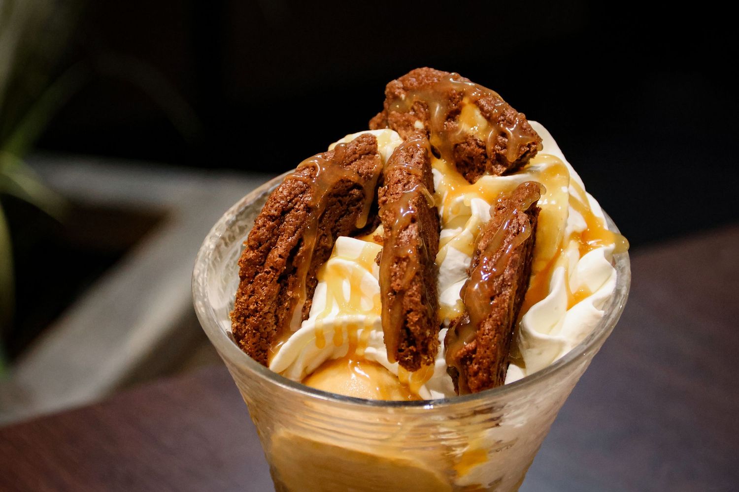 A glass dessert cup filled with vanilla ice cream, caramel sauce, and chocolate cookie pieces on a wooden table.