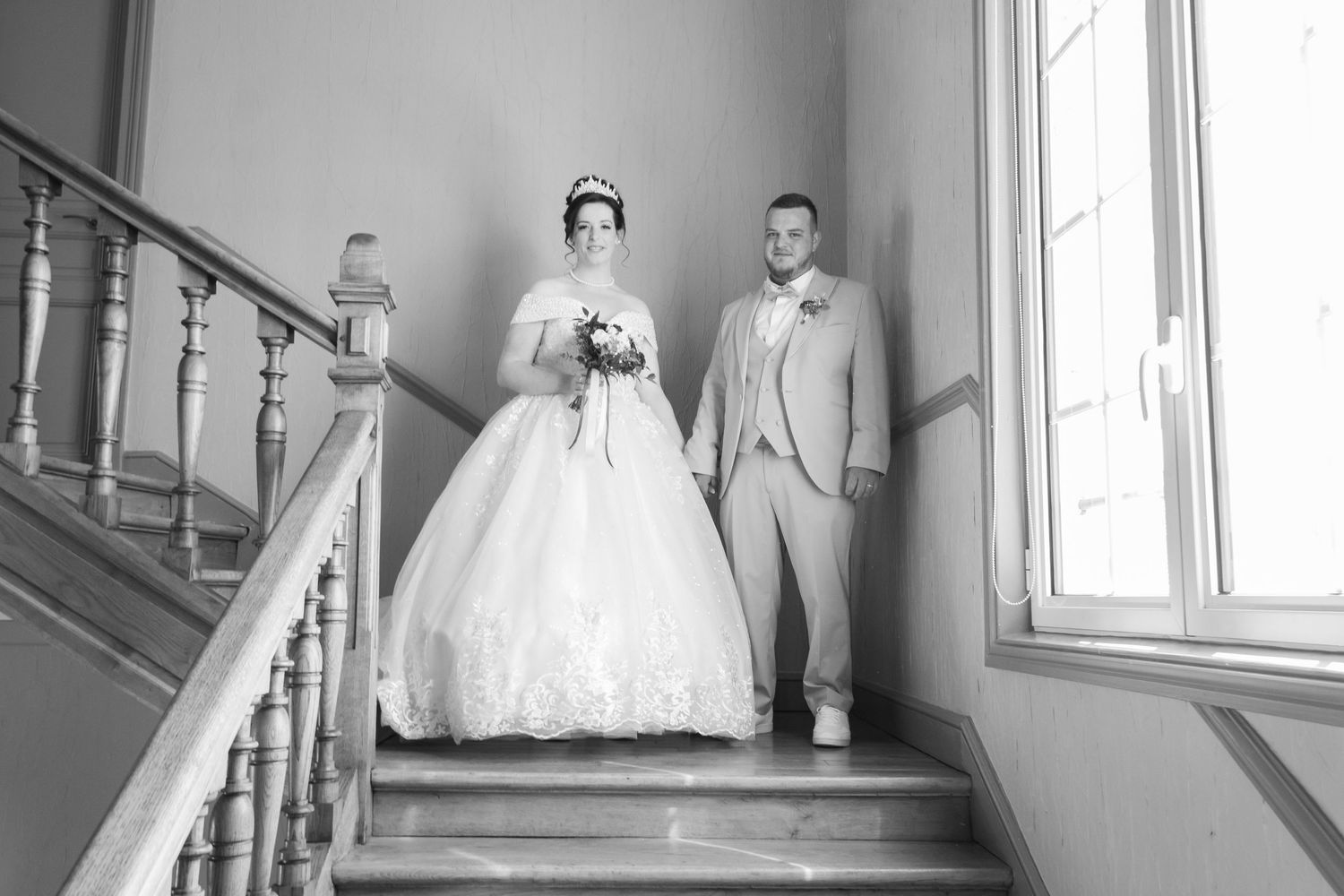 Black and white photo of a bride in a ball gown and groom in a suit posing on an elegant staircase.
