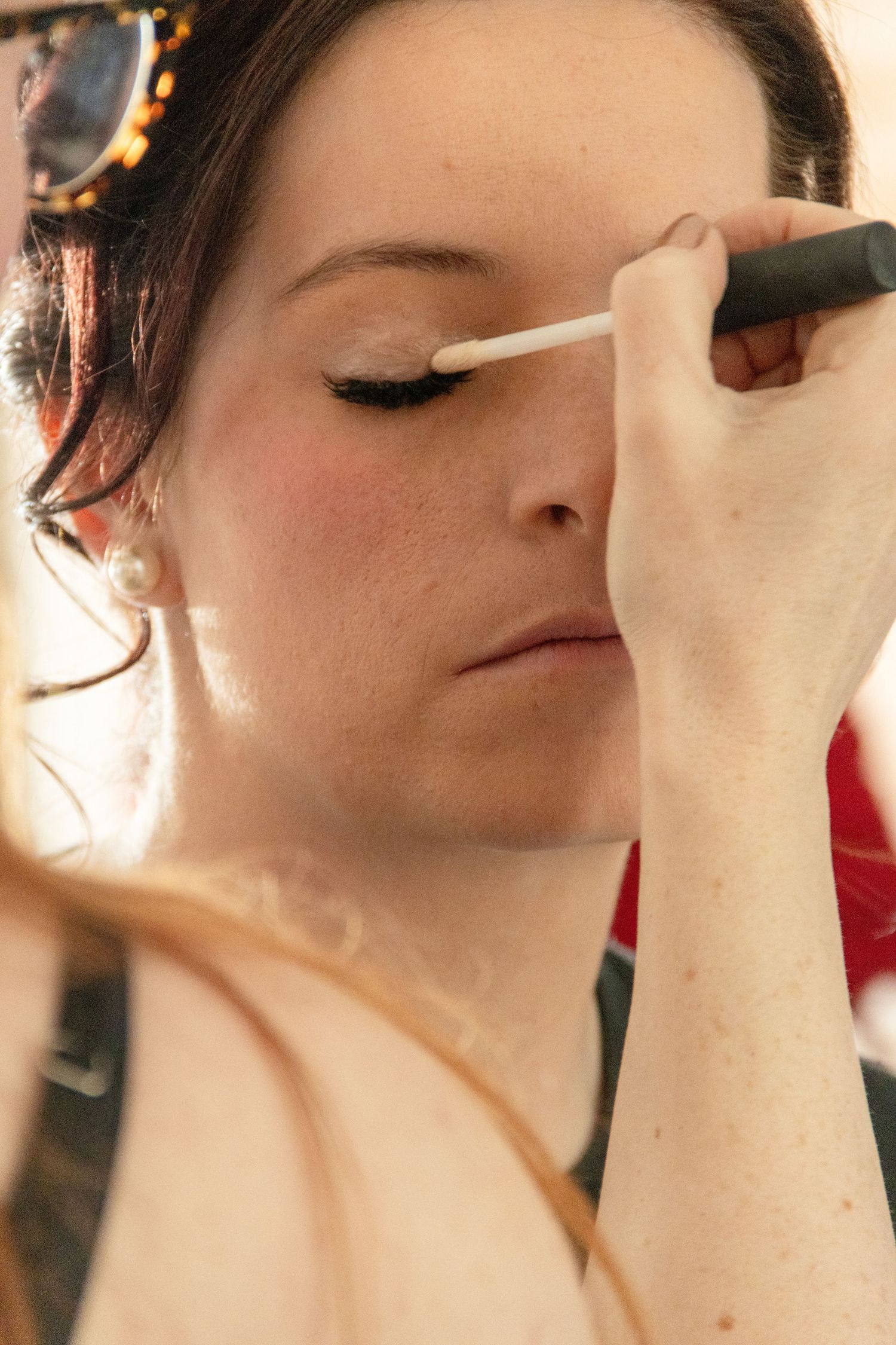 A makeup artist applying white eyeliner to a young woman wearing pearl earrings and dramatic eye makeup.