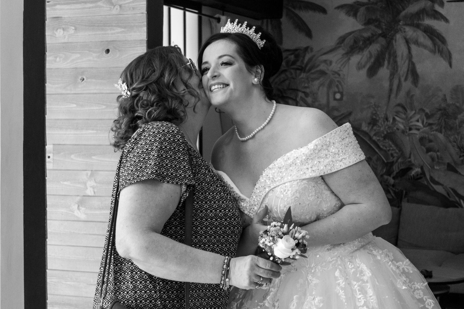 Black and white photo of a bride in tiara and off-shoulder gown sharing a joyful moment with a woman at her wedding.