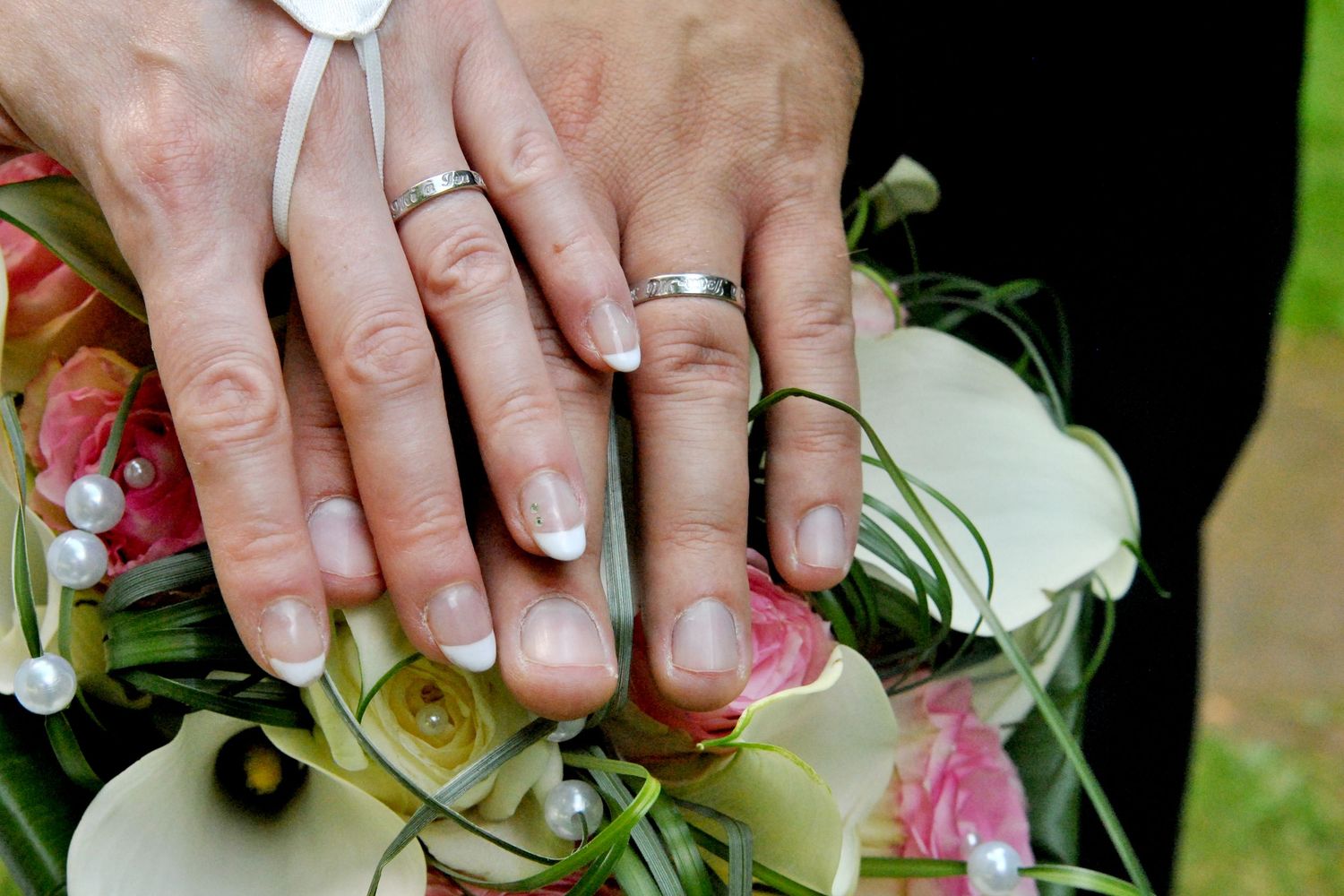 Newlyweds display their wedding rings on hands resting over a colorful bridal bouquet of white and pink flowers.