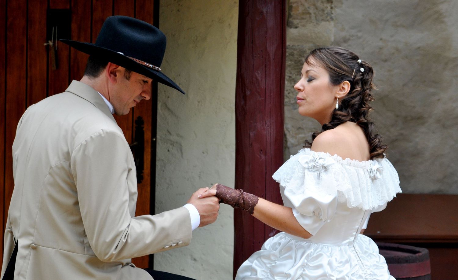 Groom in cowboy hat and tan jacket holds bride's hand in white off-shoulder wedding gown outdoors.