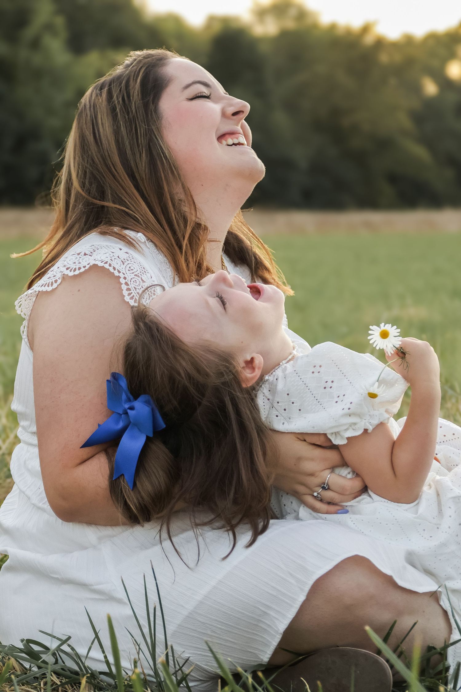 A laughing mother in white dress cuddles her young daughter holding a daisy in a sunny green field.