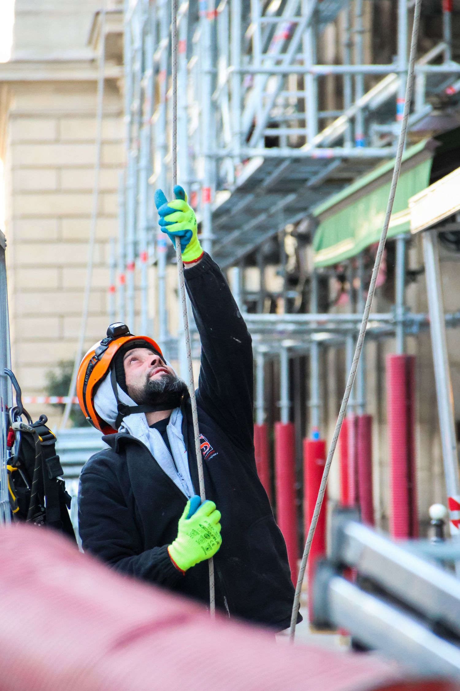 Construction worker in orange helmet and green gloves reaching up while working on scaffolding at a building site.