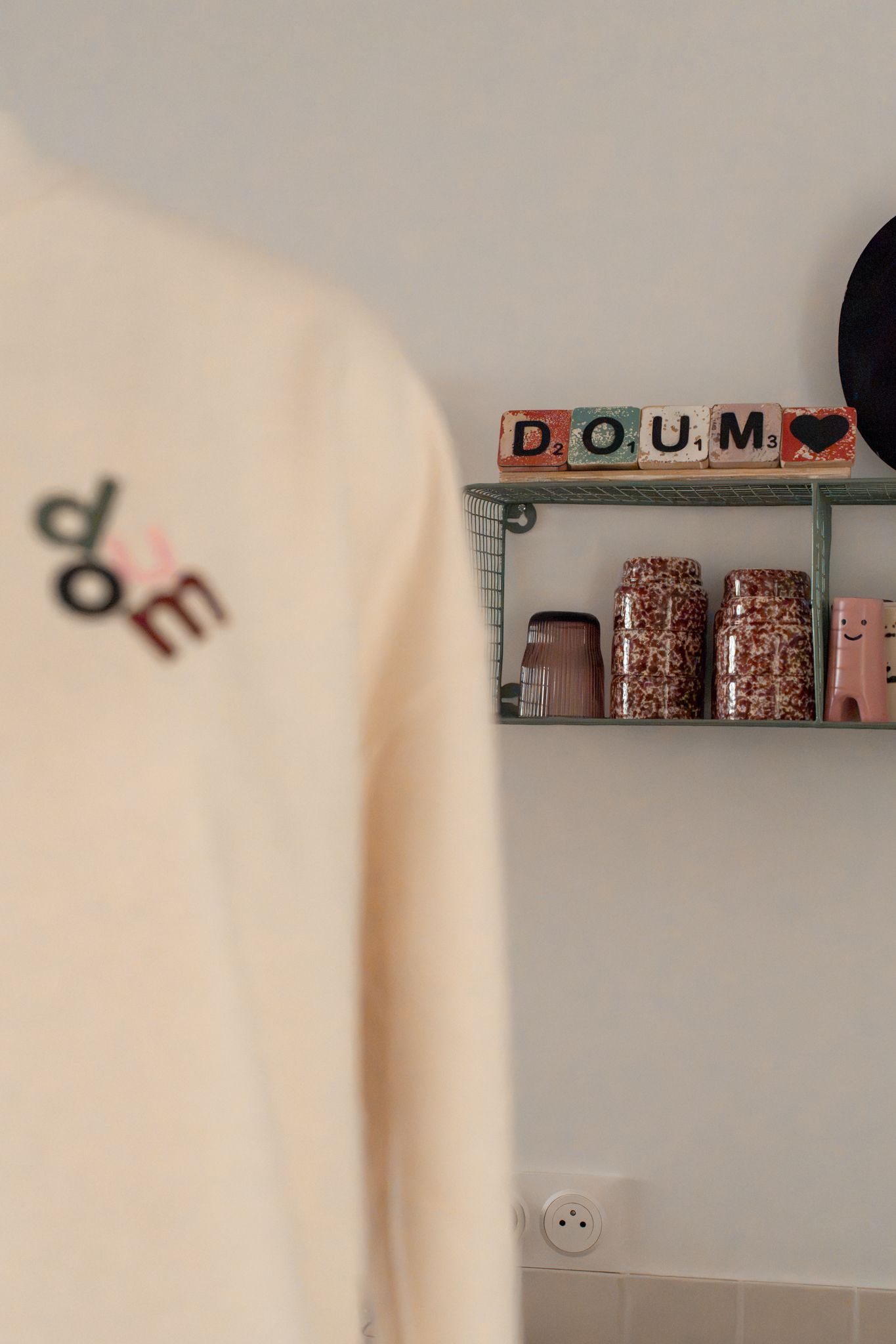 A cream sweatshirt with 'doe' embroidery beside a shelf displaying 'D.O.U.M' Scrabble tiles and decorative jars.