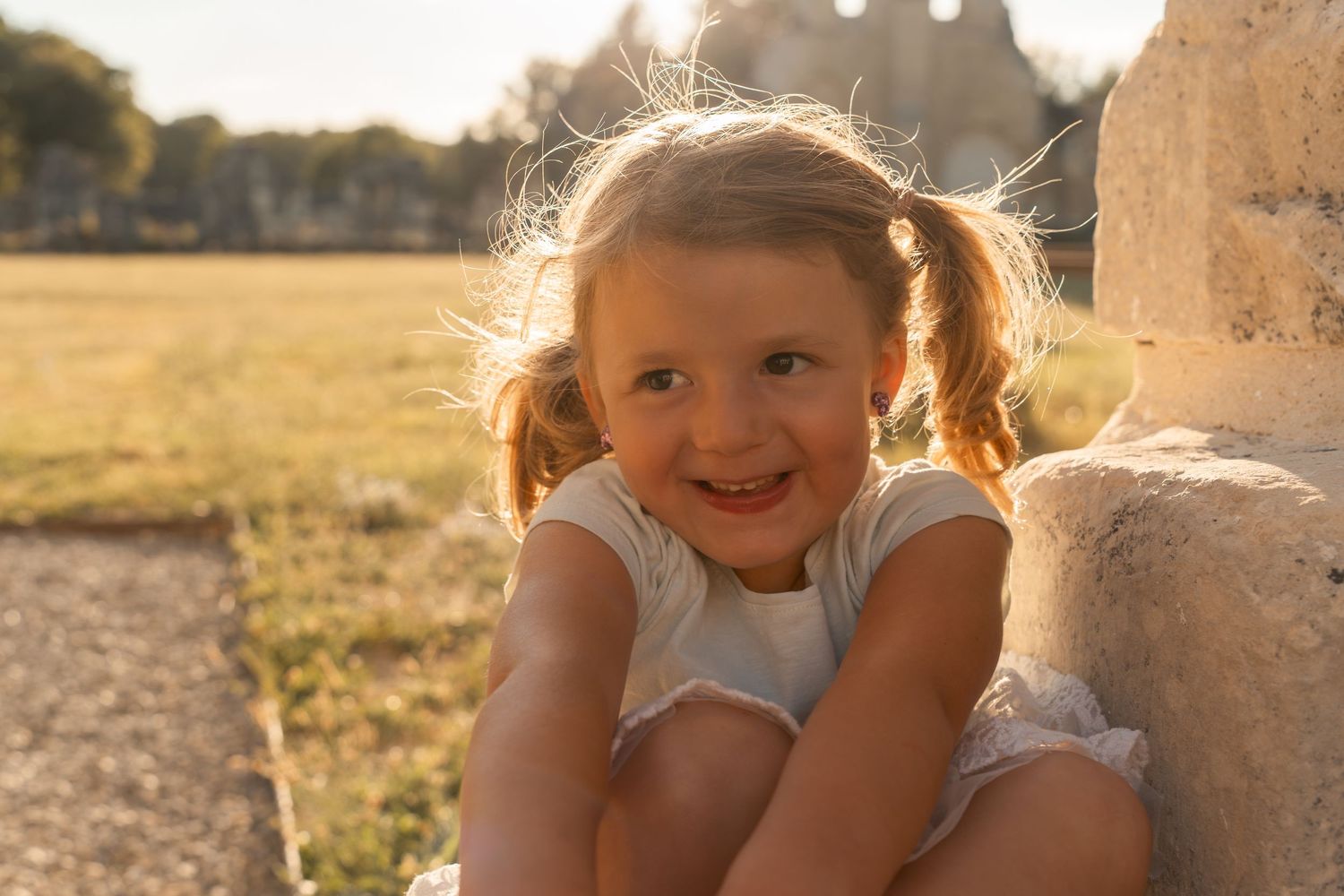 Adorable smiling little girl with pigtails sitting against a stone wall in golden afternoon sunlight.