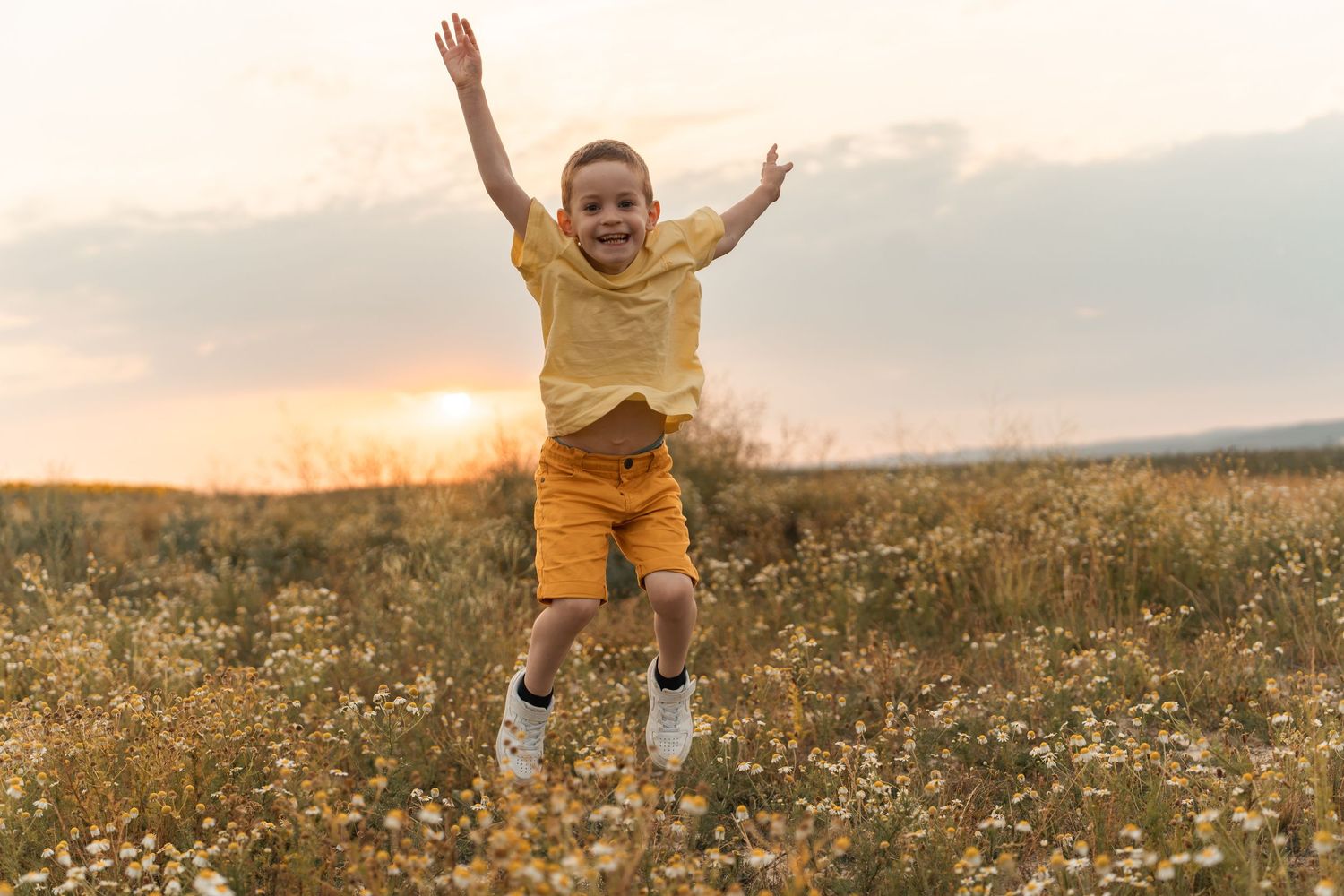 Joyful young boy in yellow outfit jumping in a flower field at sunset with arms raised.