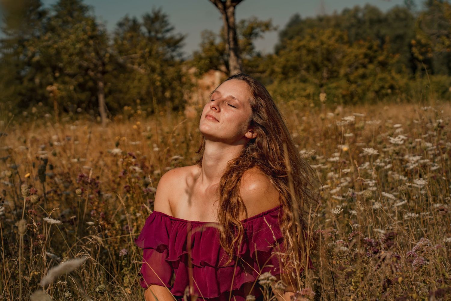 Young woman with long hair in red off-shoulder dress sitting in a wildflower meadow, eyes closed, basking in sunlight.