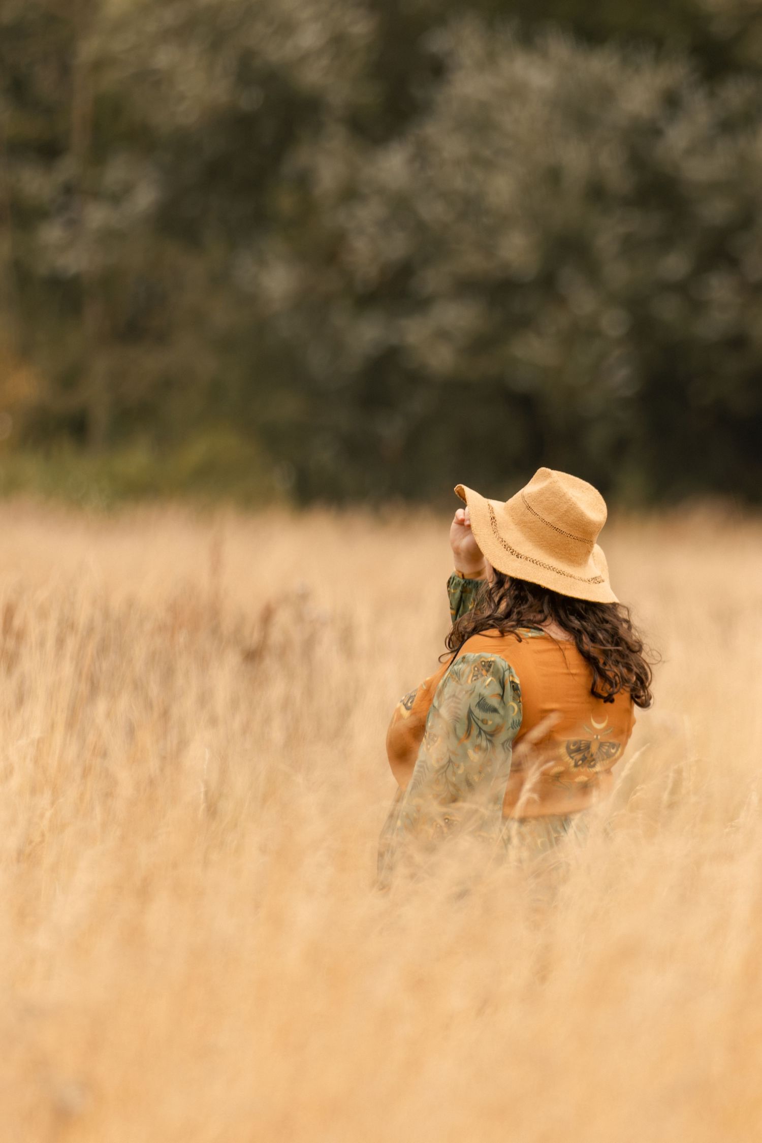 Woman in straw hat and orange jacket standing in golden wheat field with blurred green trees background.