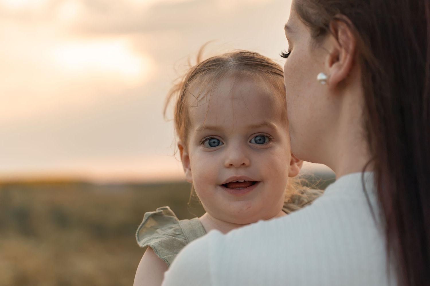 A smiling baby with blue eyes being held by a mother outdoors at sunset.