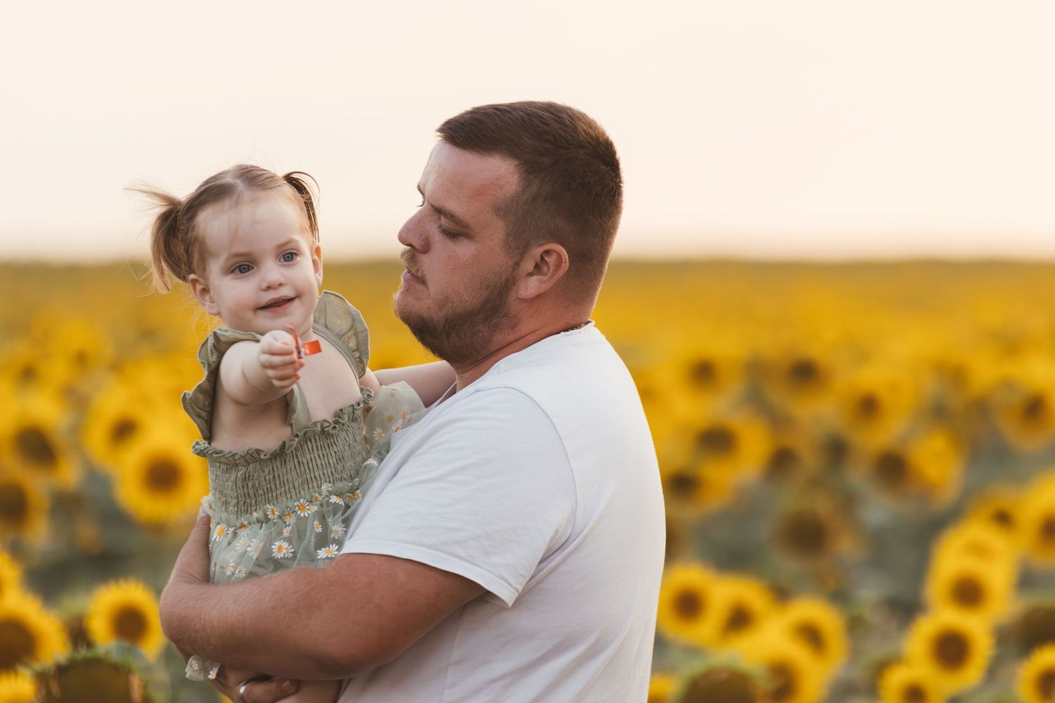A father holds his toddler daughter in a sunflower field at golden hour.