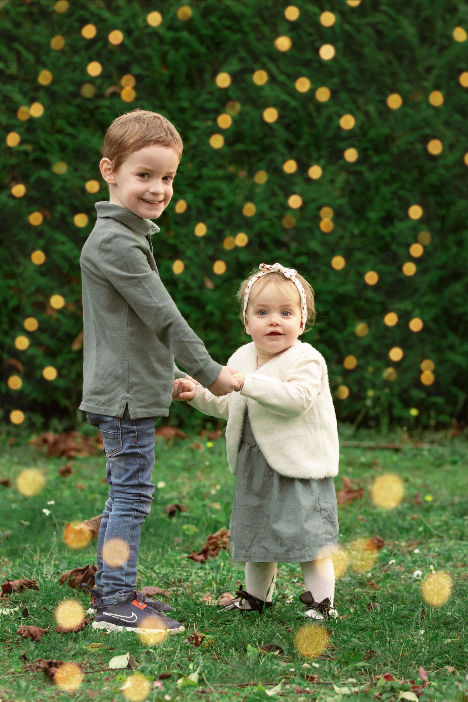 A young boy holds hands with a baby girl on green grass with golden bokeh lights.