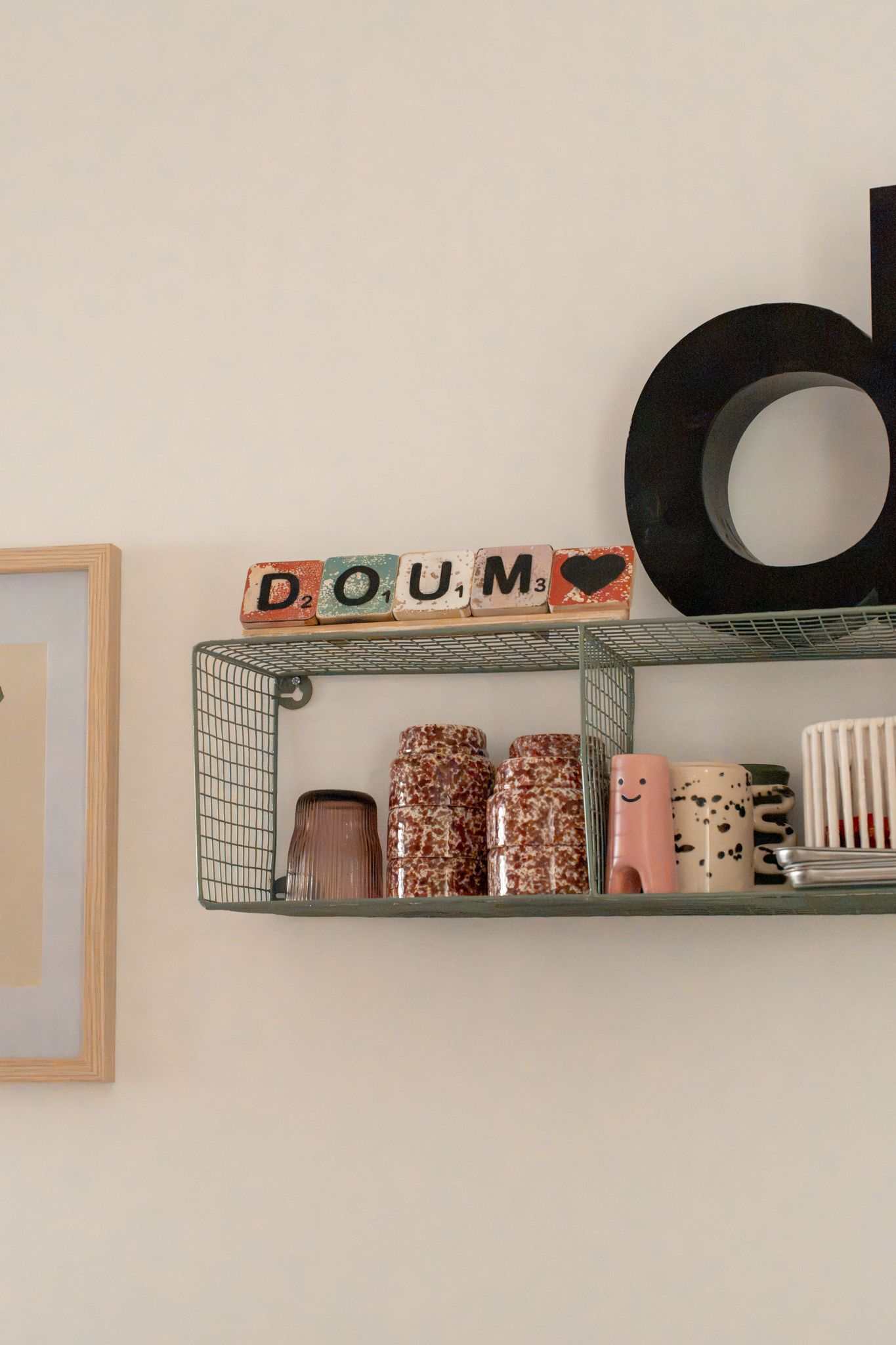 Green wire shelf displaying Scrabble tiles spelling DOUM, ceramic mugs, and a black letter D decorative object on a white wall.
