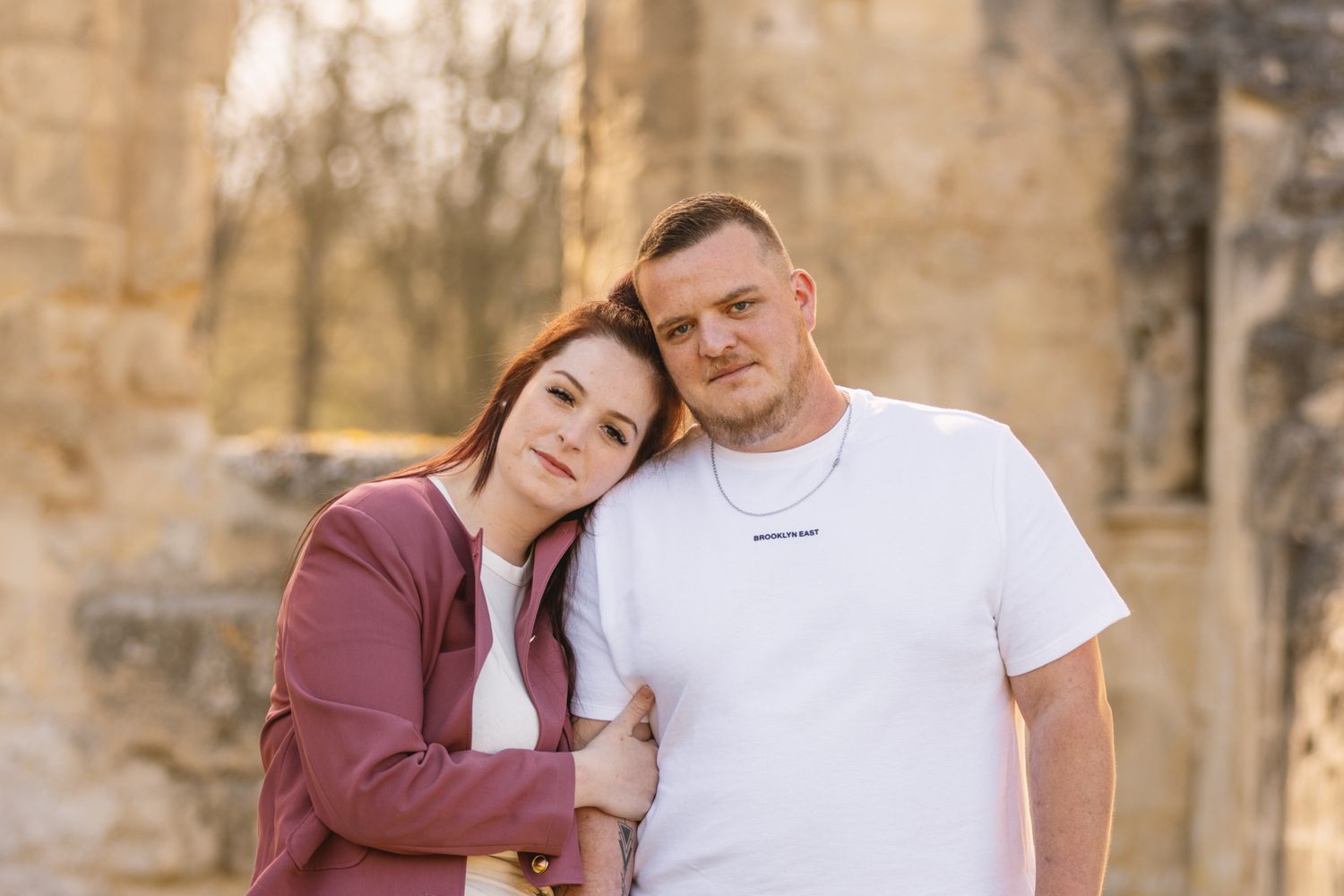 A couple poses together outdoors, woman in pink jacket leaning on mans shoulder in white tee, stone ruins background.