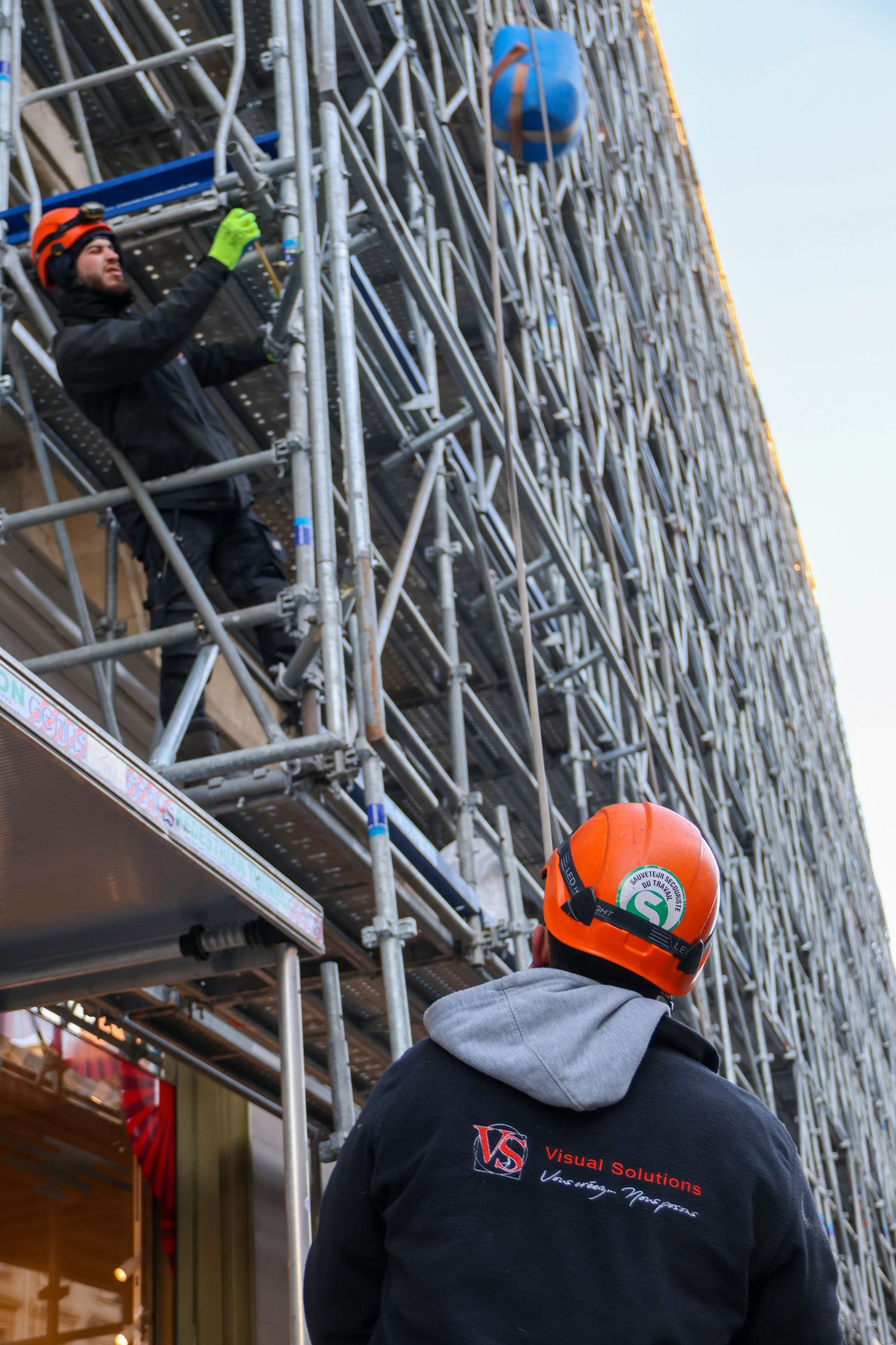 Two construction workers in orange hard hats work on metal scaffolding outside a building, one tossing a blue ball.