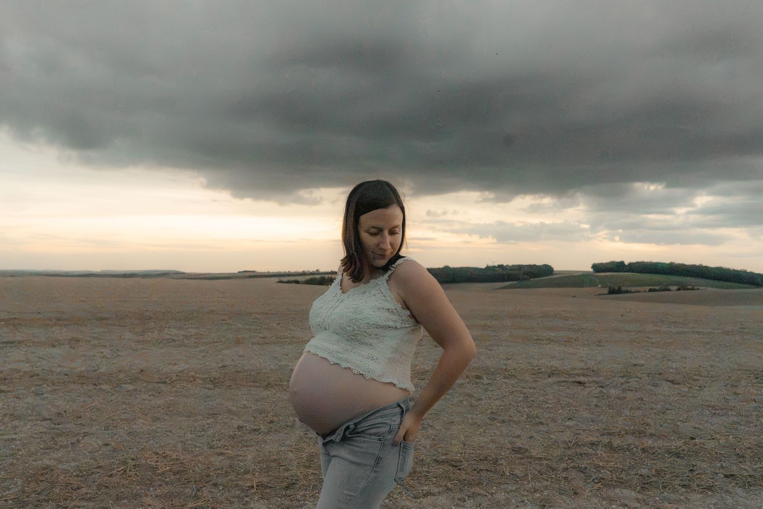 Pregnant woman in white top and jeans stands in open field under dramatic cloudy sky at dusk.