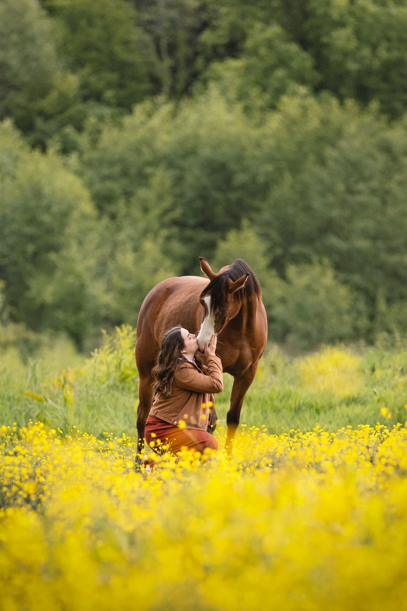 Woman kneeling in yellow wildflower field bonding with a brown horse, surrounded by lush green trees.