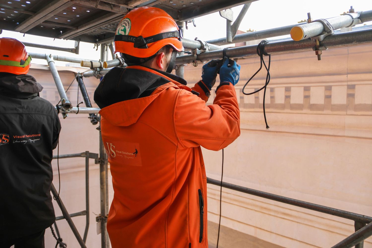 Workers in orange safety gear and helmets perform maintenance on industrial equipment on an elevated platform.