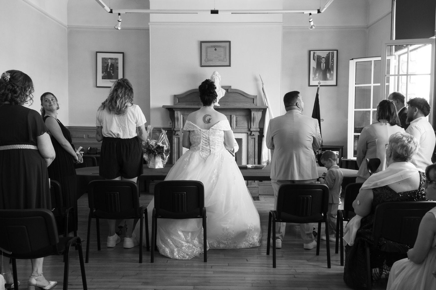 Black and white wedding ceremony photo showing bride and groom at altar with guests seated in chairs inside a hall.