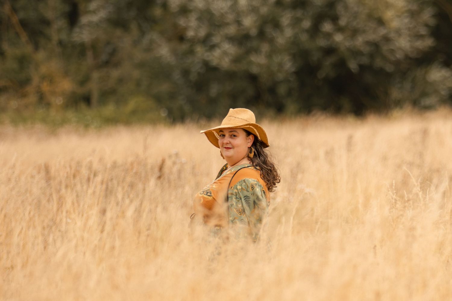 Woman in yellow cowboy hat smiling in golden wheat field with green trees in background.
