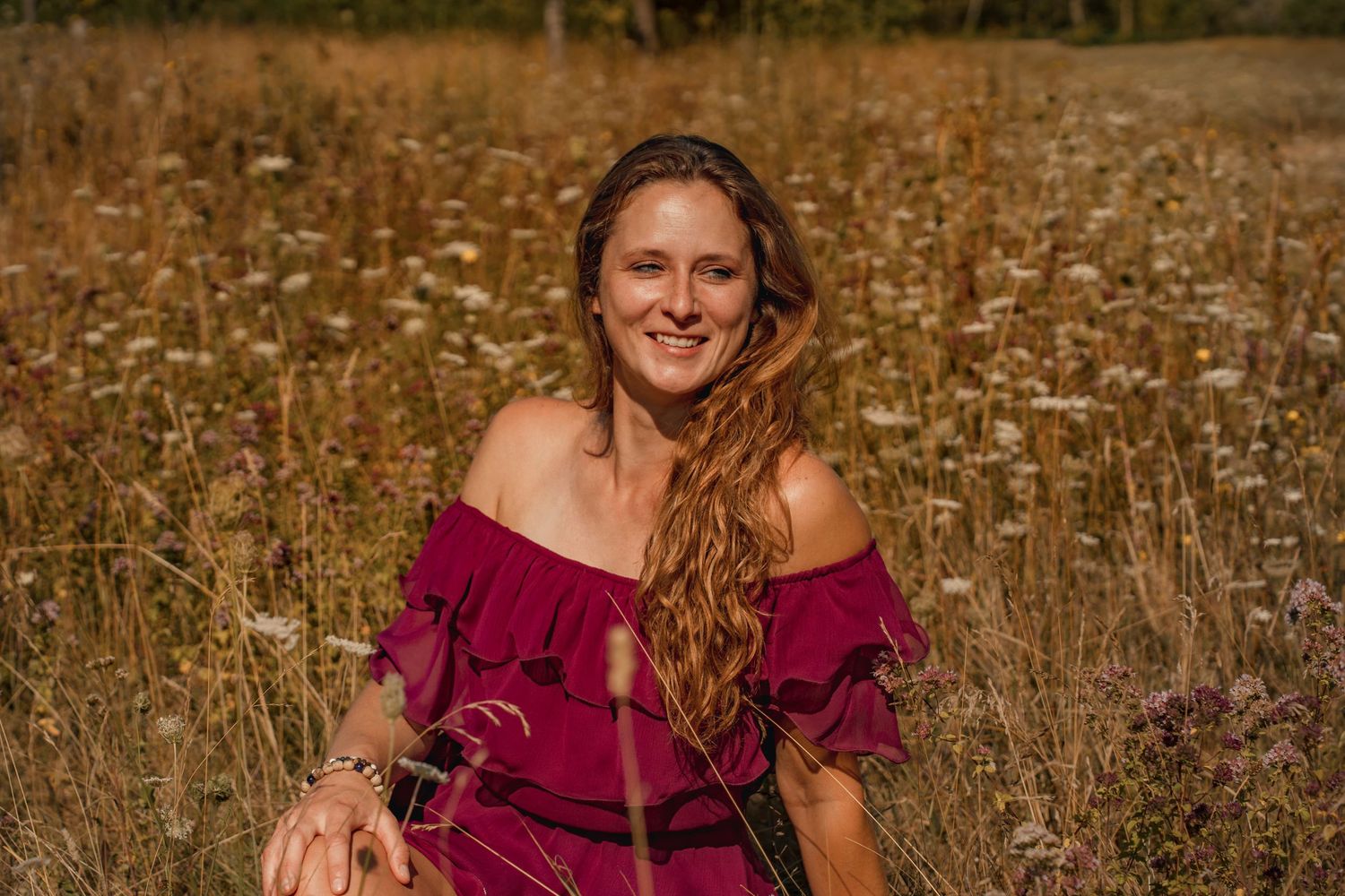Smiling woman with long wavy hair in red off-shoulder dress sitting in a sunny wildflower meadow.