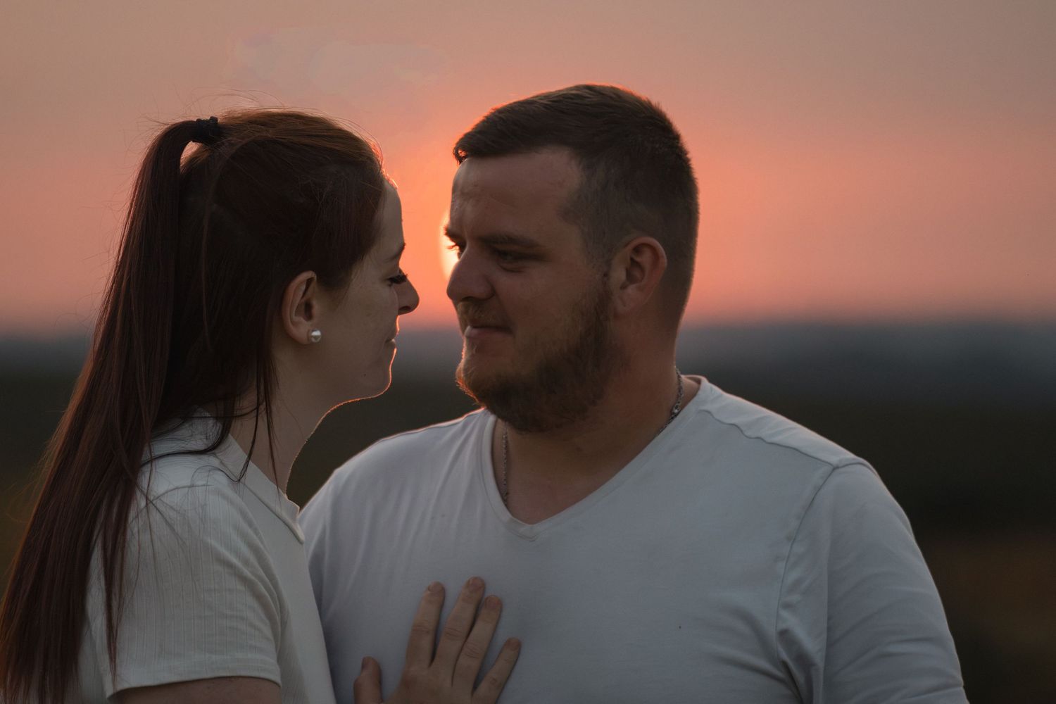 Romantic couple gazing at each other during a stunning sunset, wearing white shirts in an outdoor setting.