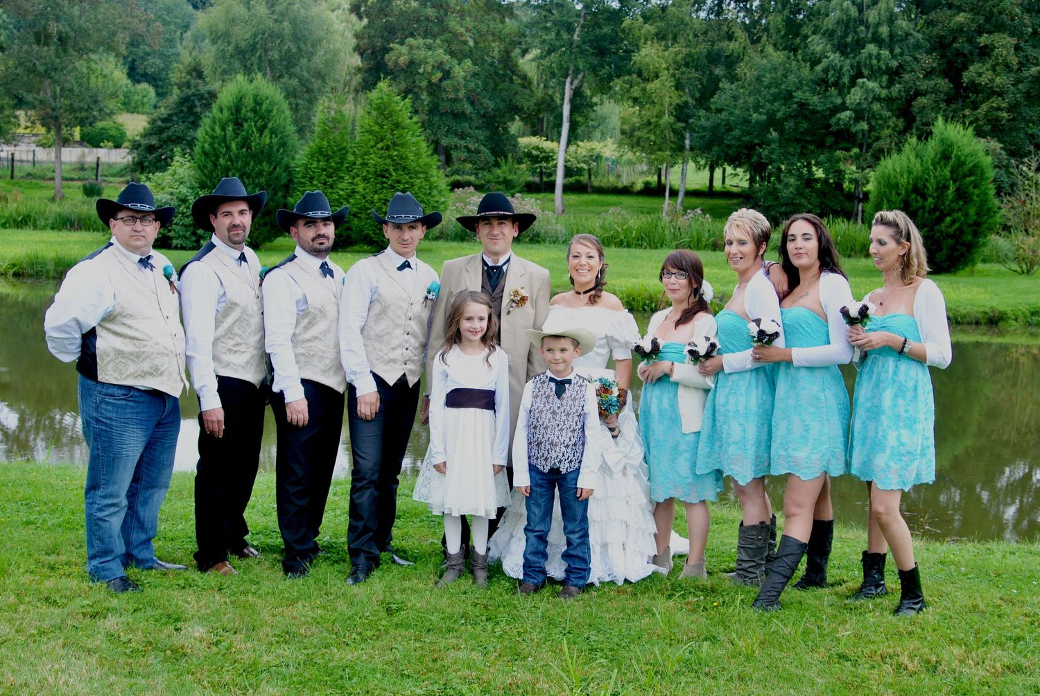 Western-themed wedding party posing near a pond with groomsmen in cowboy hats and bridesmaids in teal lace dresses.