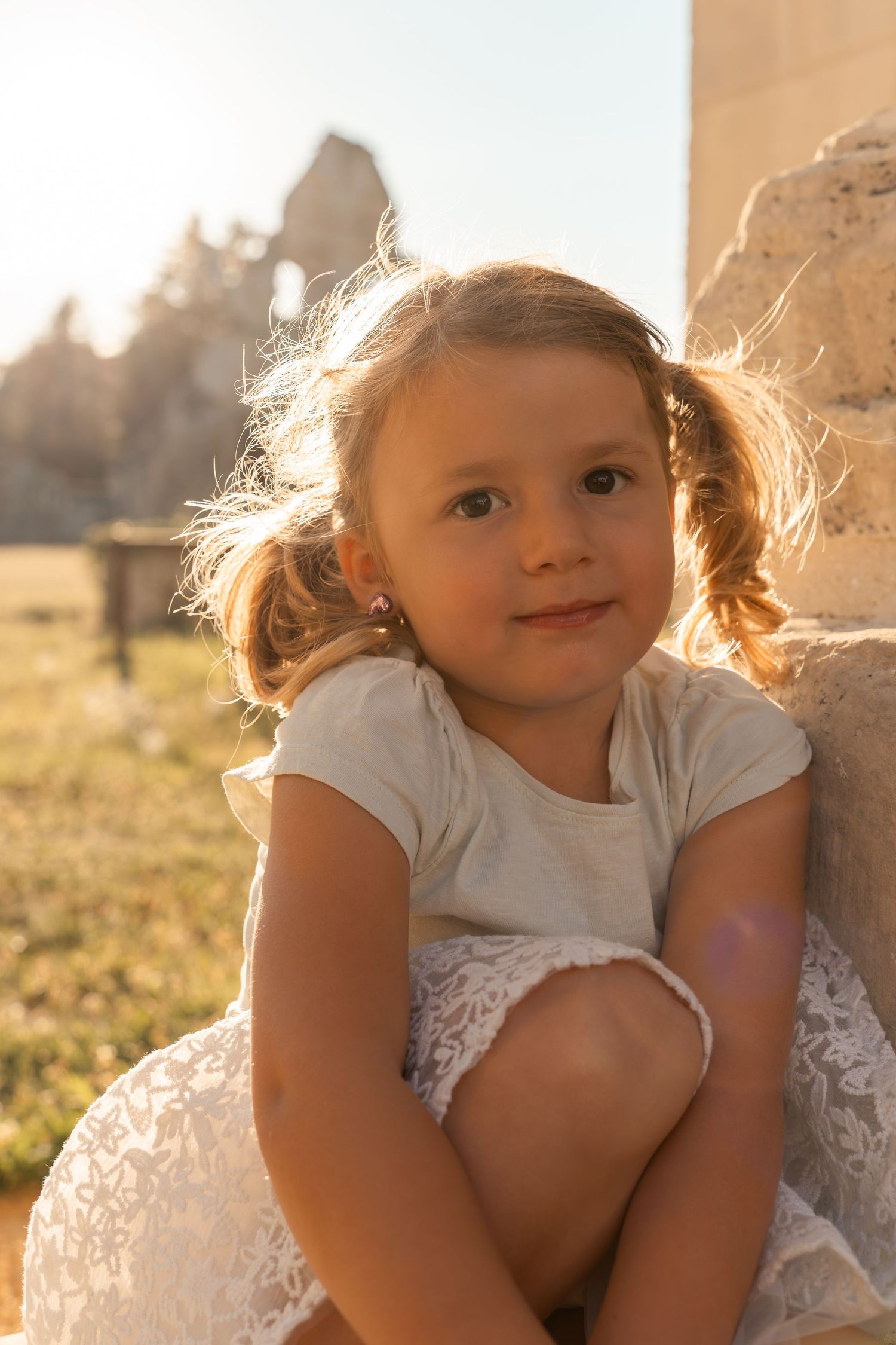 A cute little girl with pigtails in a white dress sits peacefully by a stone wall at sunset.