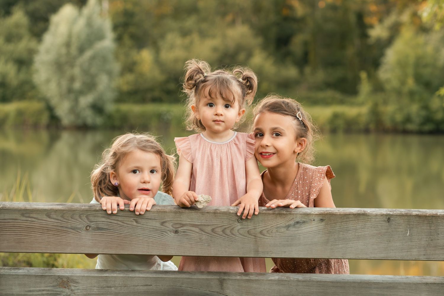 Three young sisters in pink dresses peek over a wooden fence near a peaceful lake outdoors.