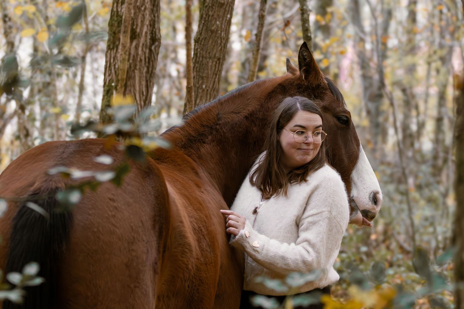 Smiling woman in white sweater standing beside a chestnut horse among autumn trees.