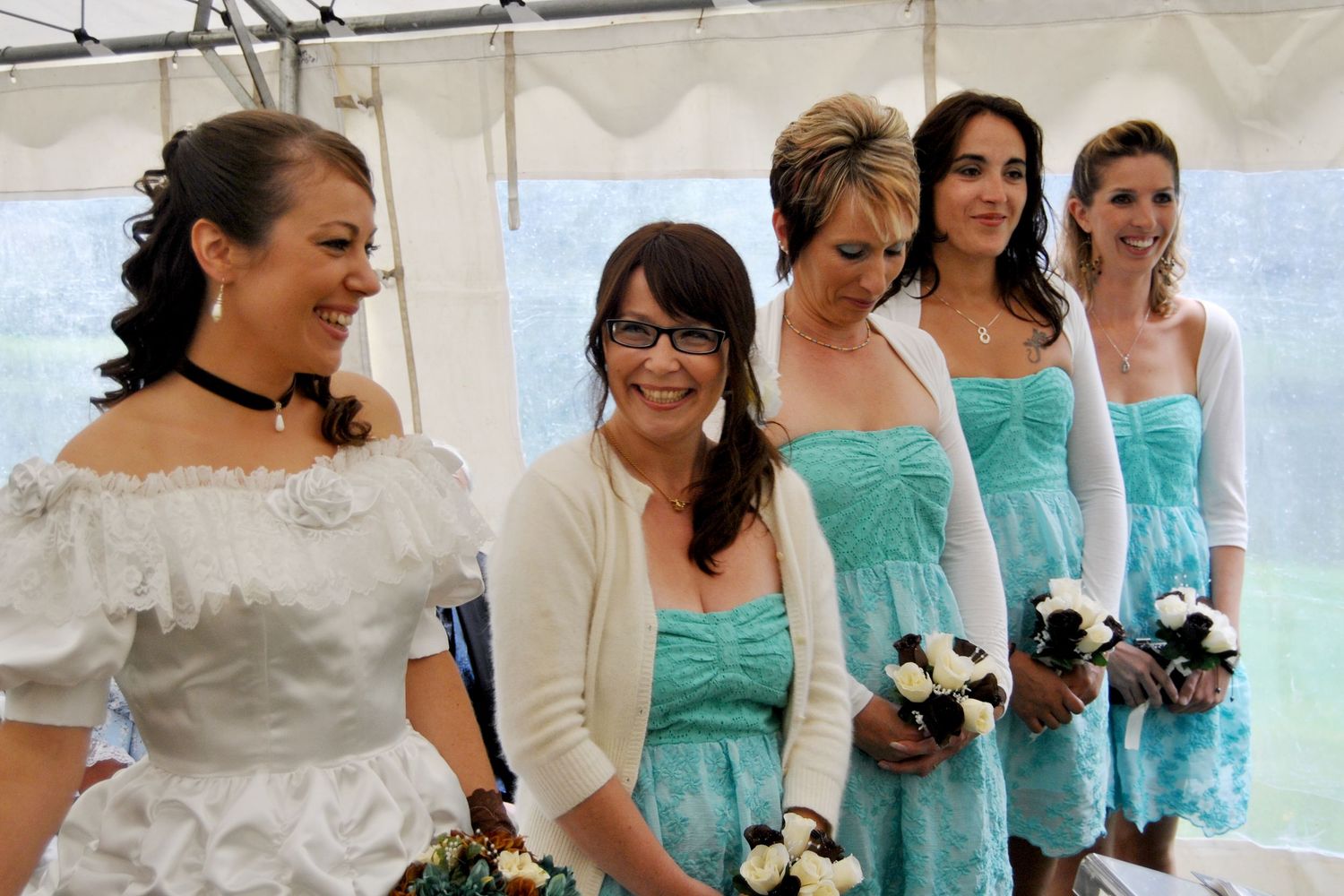 Bride in white ruffled gown poses with four bridesmaids in teal dresses holding white bouquets at a western wedding.