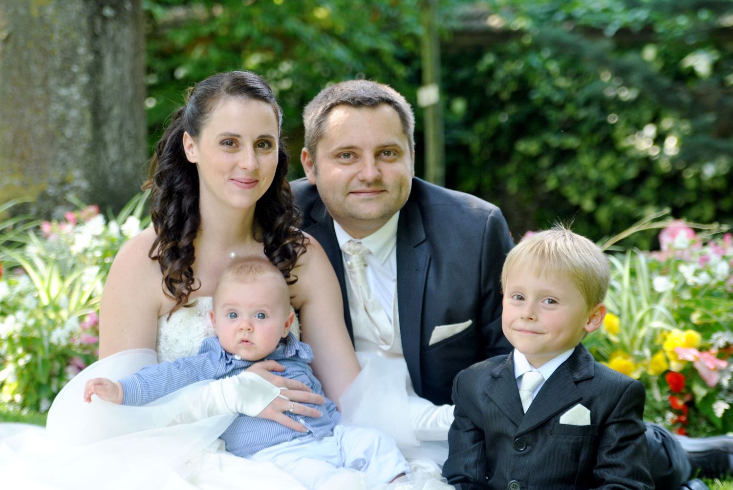 Wedding couple smiles with two young children in colorful garden setting.