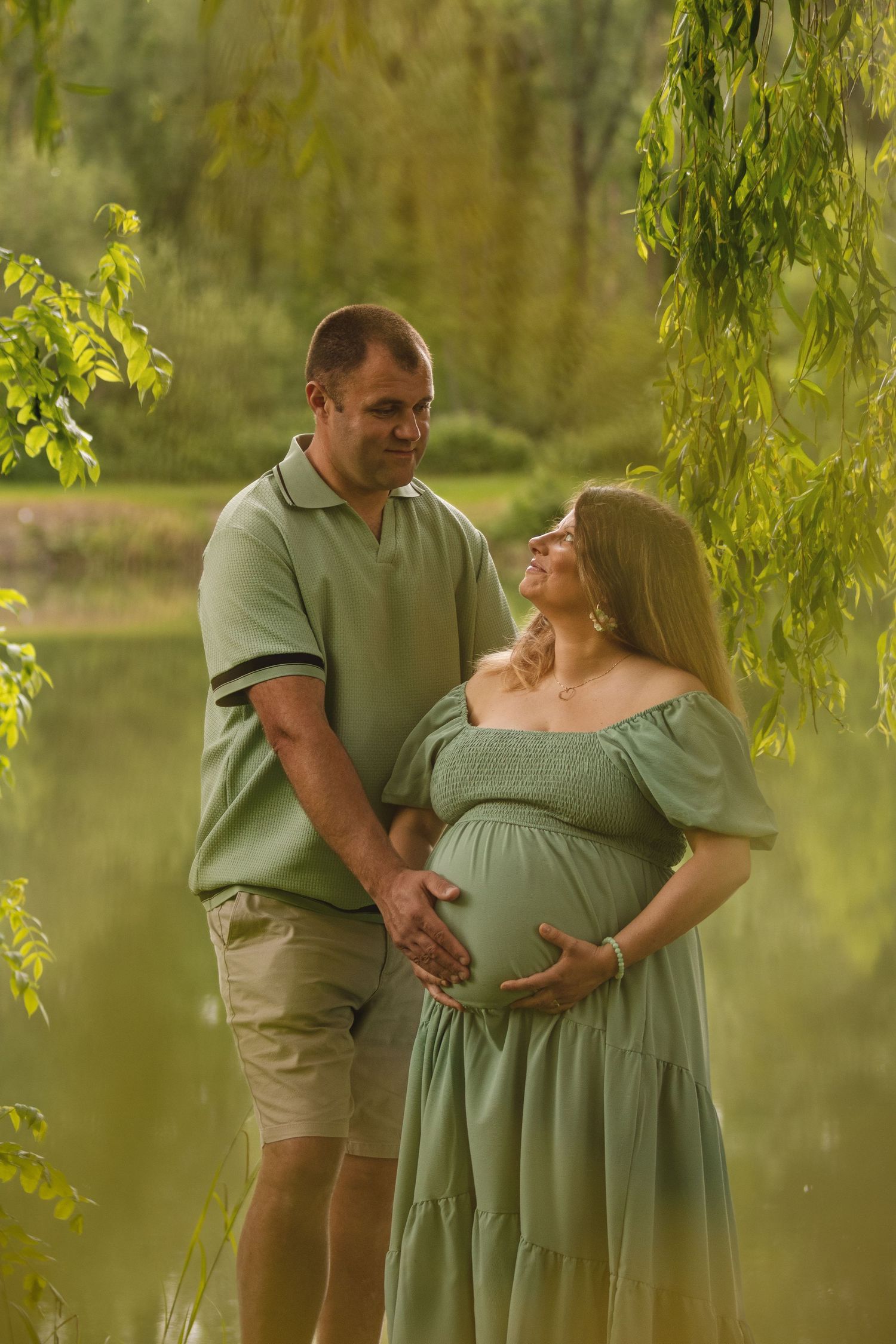 A couple in matching green outfits tenderly cradle a pregnant belly outdoors near a serene pond with lush greenery.